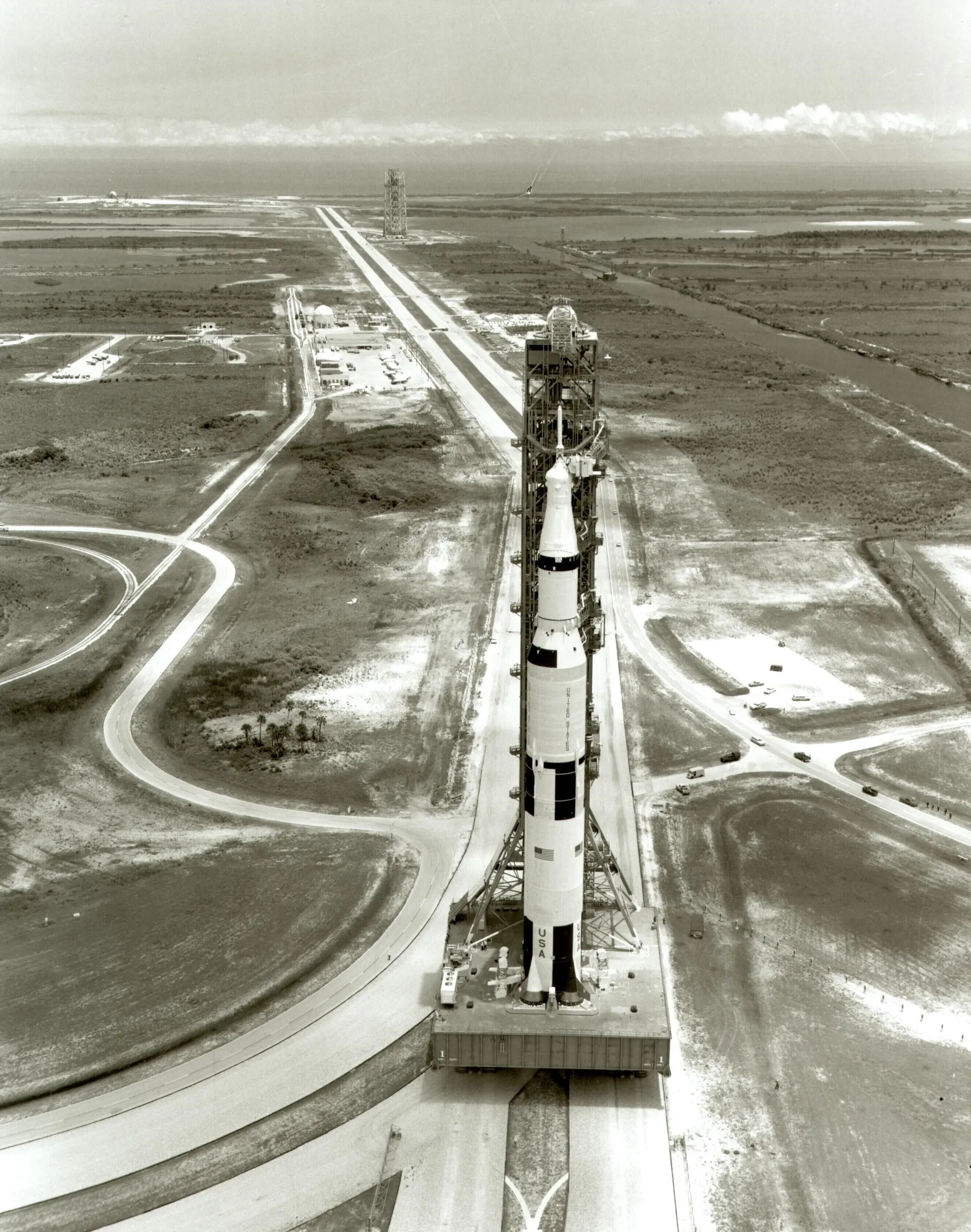 Seen from the air, the Apollo 11 Saturn V rides its crawler up the crawlerway toward Pad 39A, with the ocean on the horizon.