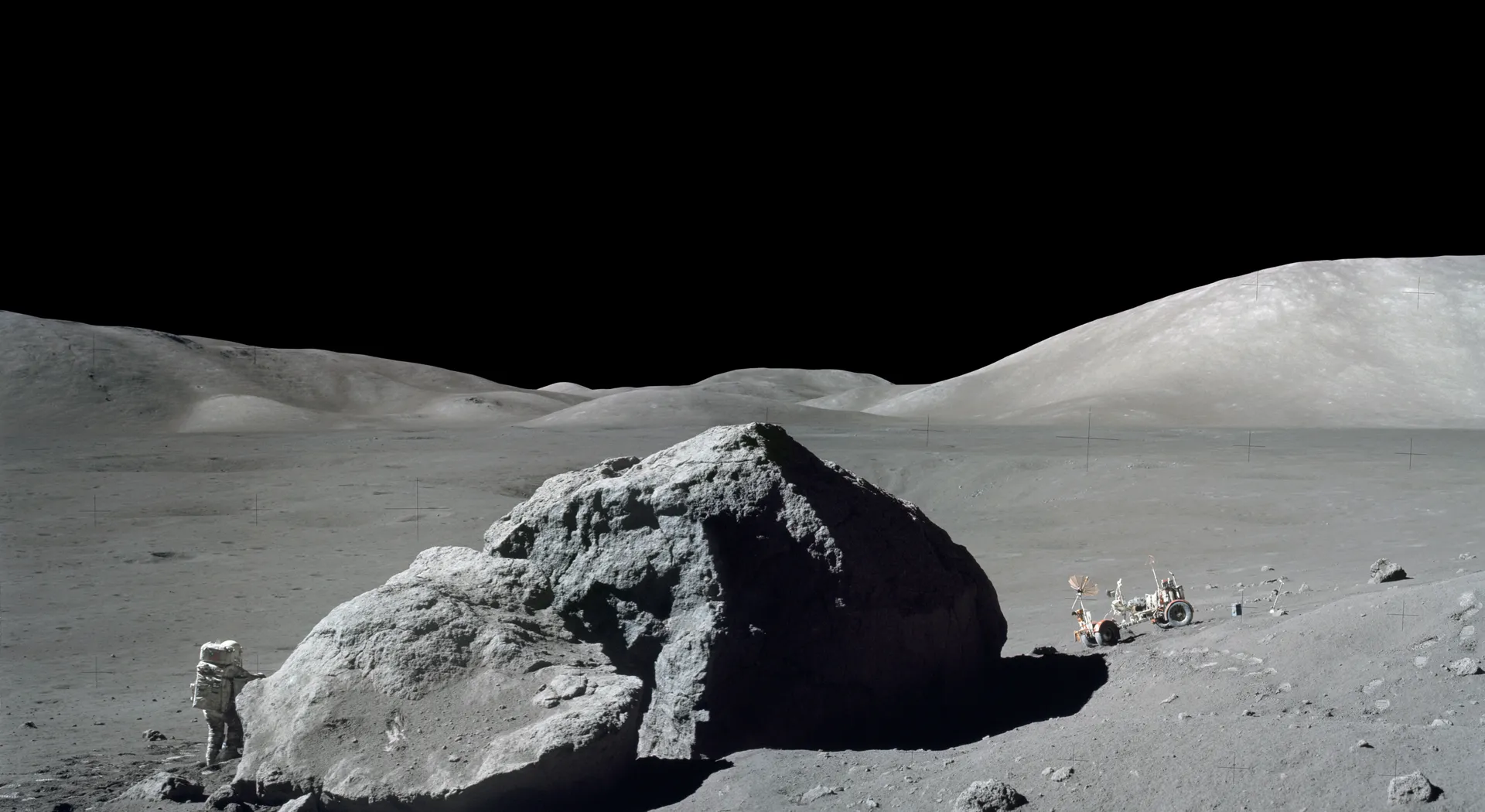 Harrison Schmitt, small beside a huge split boulder, with the rover parked to the right and mountains under a black sky.