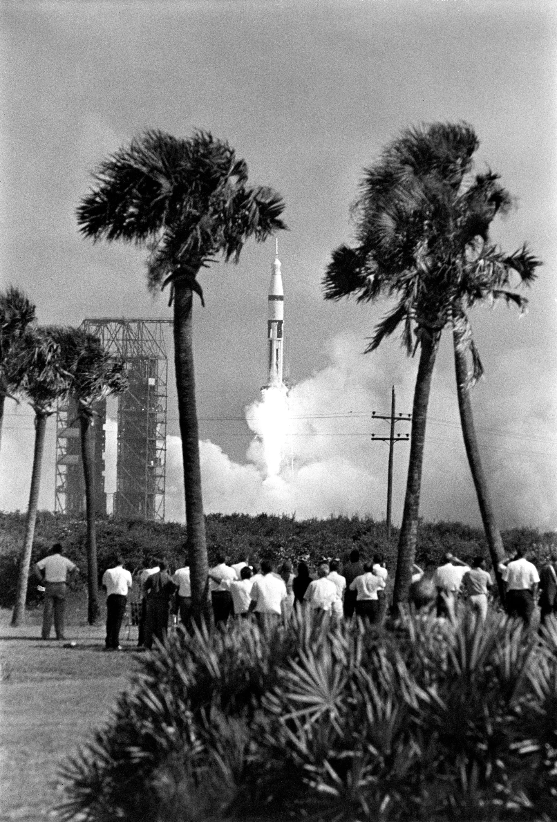 A Saturn IB rises from its pad beyond palm trees as spectators watch from a field.