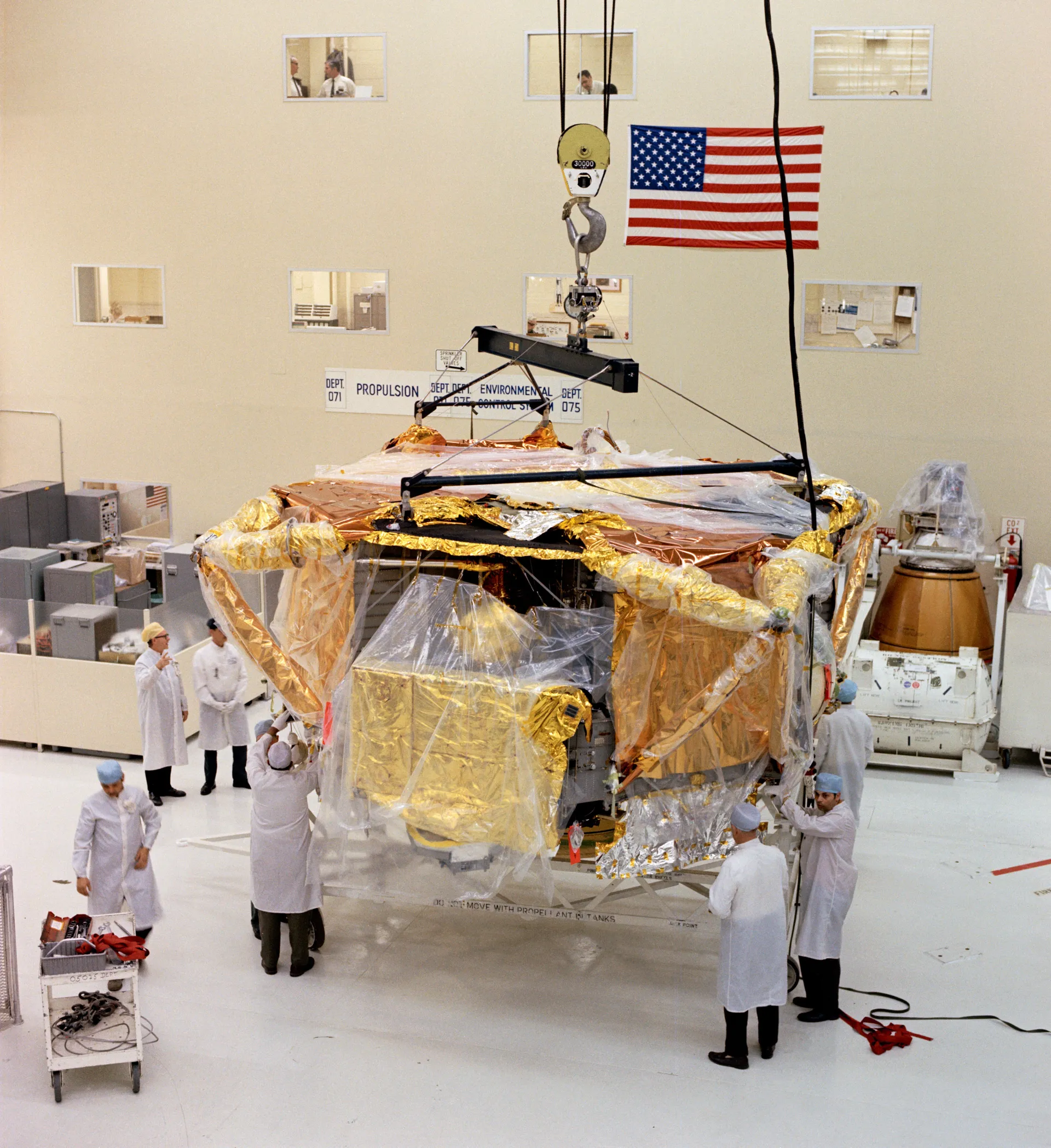 Gowned technicians guide a foil-wrapped lunar module descent stage hanging from a hoist in a clean room.