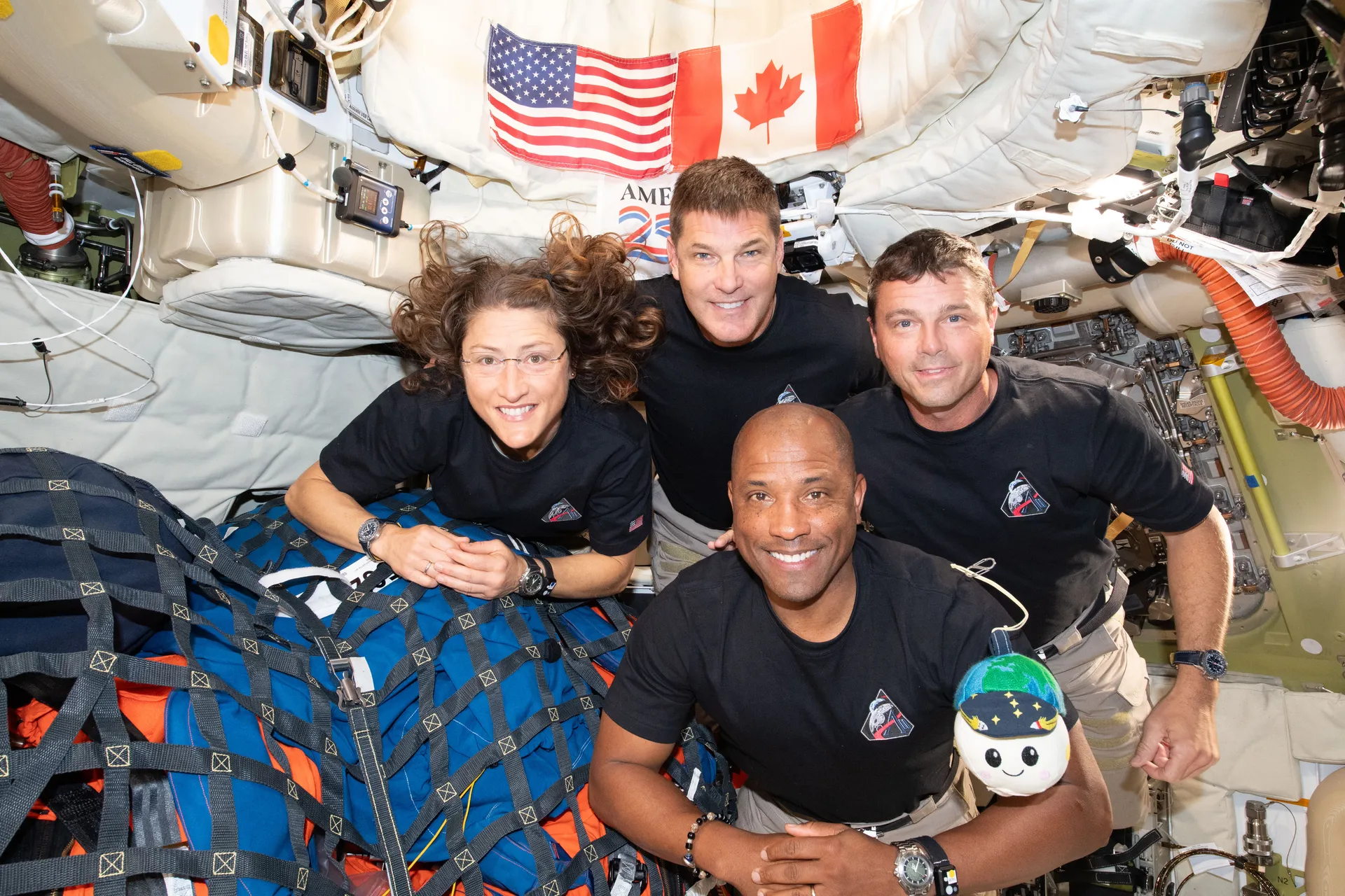 Christina Koch, Jeremy Hansen, Reid Wiseman and Victor Glover smile at the camera inside Orion’s cabin, with U.S. and Canadian flags behind them and a small plush toy floating in front.