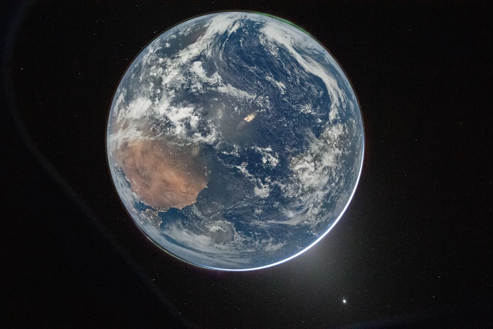 The full disk of Earth, with Africa at left under swirling clouds and faint green auroras at its edges, seen through an Orion window against black space.