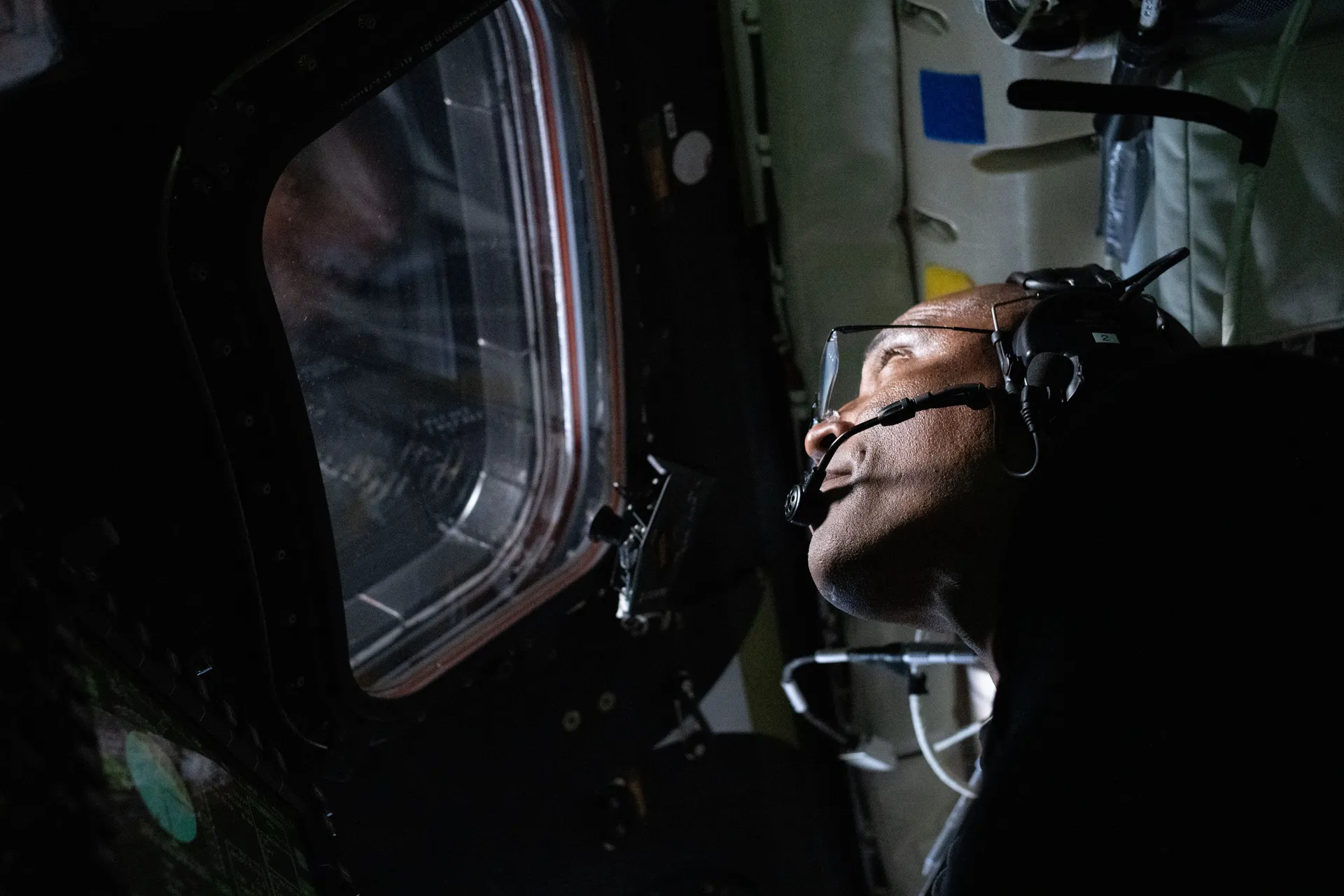 Victor Glover, wearing a headset, looks up and out of an Orion window in the dim cabin.