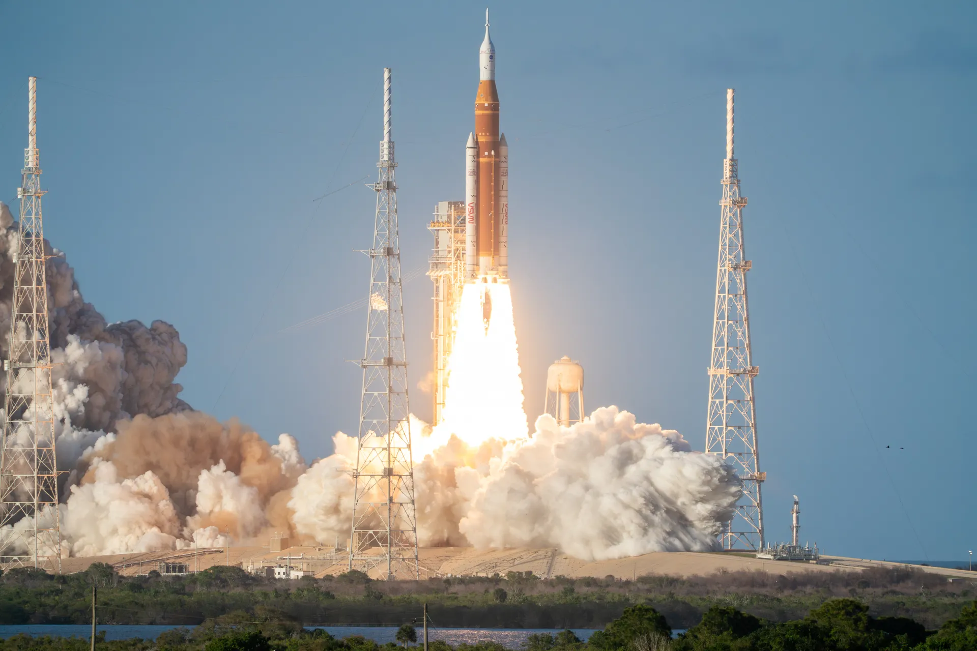 The SLS rocket rises from Launch Pad 39B on a column of flame and white exhaust, between two lightning towers and a water tower, under a clear blue sky.