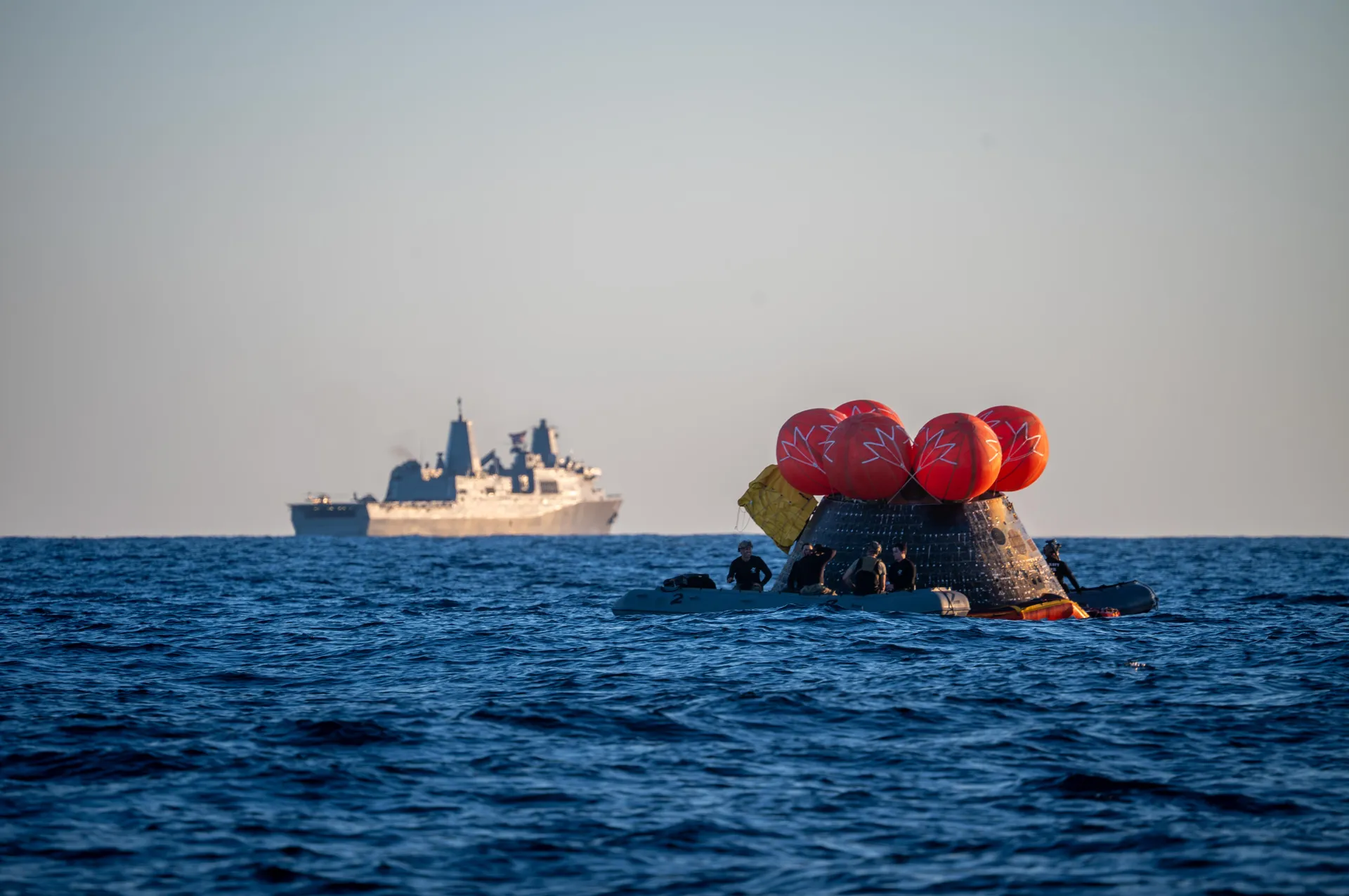Orion floats in the Pacific with orange uprighting bags inflated on top and Navy divers alongside, while the amphibious ship USS John P. Murtha waits on the horizon.