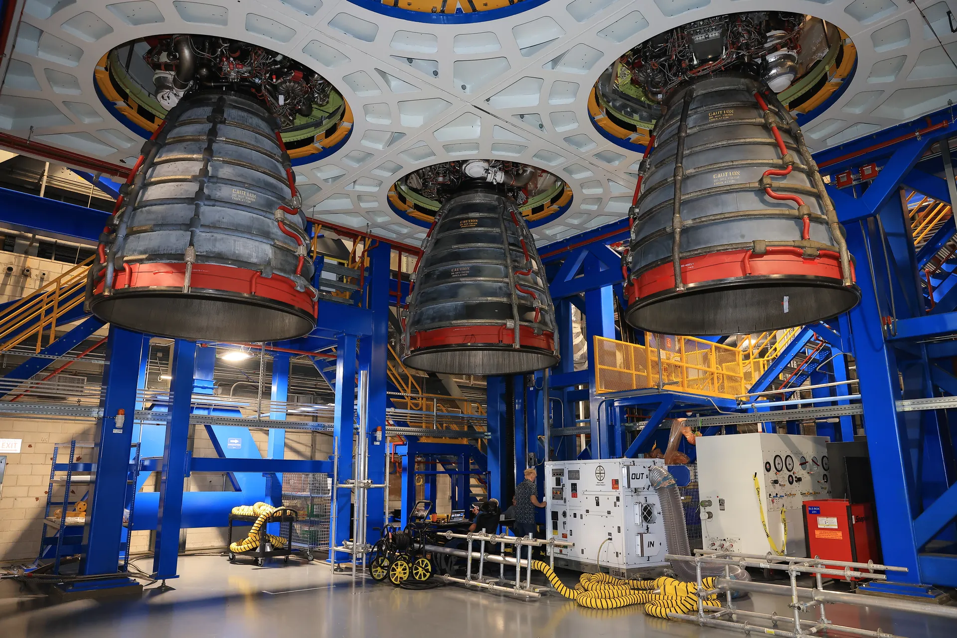 Three RS-25 engine bells hang from the base of the Artemis III core stage, seen from below inside the blue steel work platforms of the Vehicle Assembly Building.