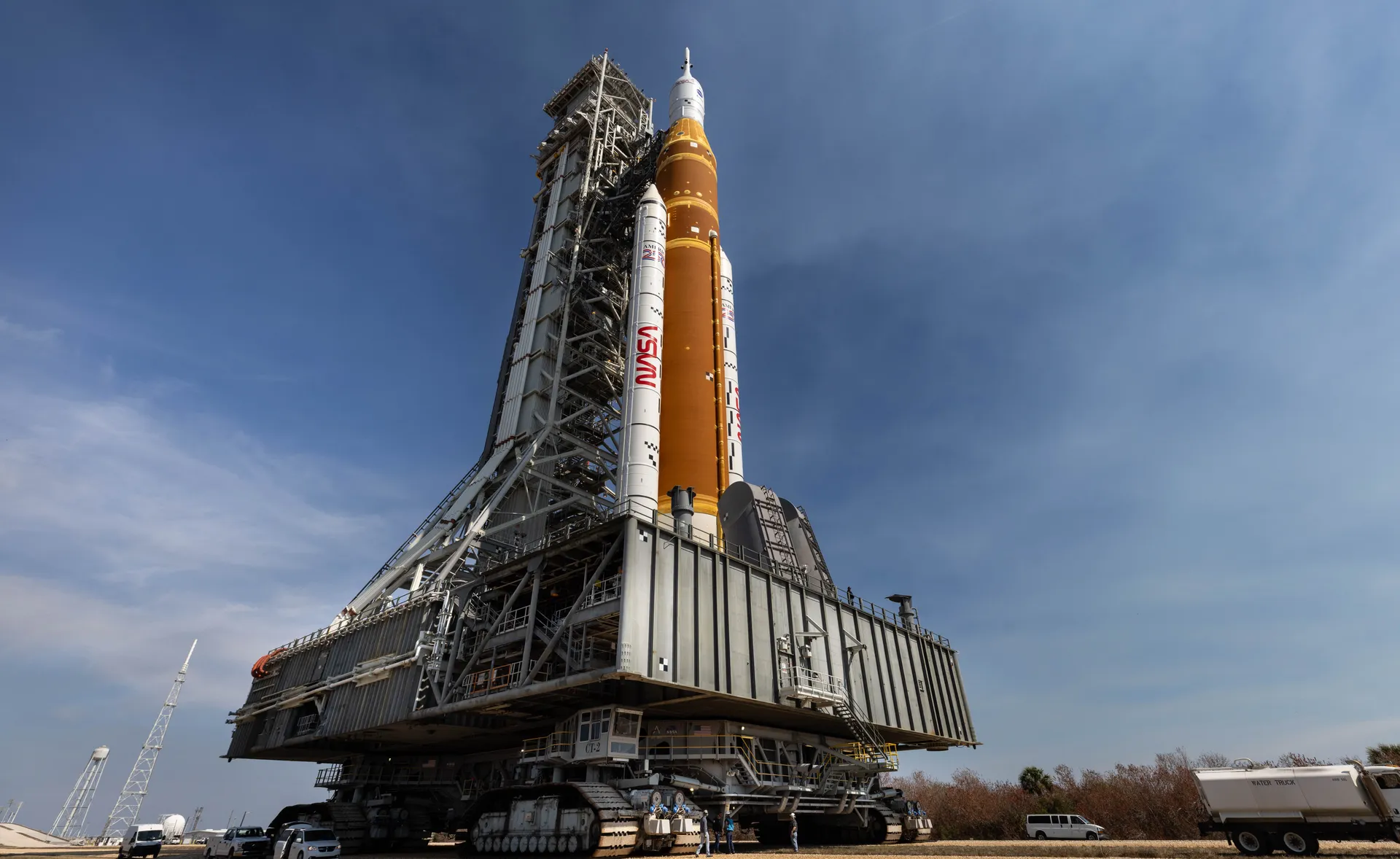 The crawler-transporter carries the Artemis II SLS rocket and its mobile launcher tower along a gravel crawlerway under a blue sky.