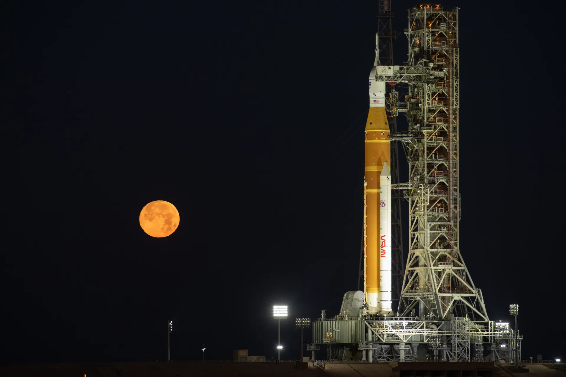 The full Moon, orange and low in a black sky, rises to the left of the Artemis II SLS rocket and Orion spacecraft standing beside their launch tower at night.