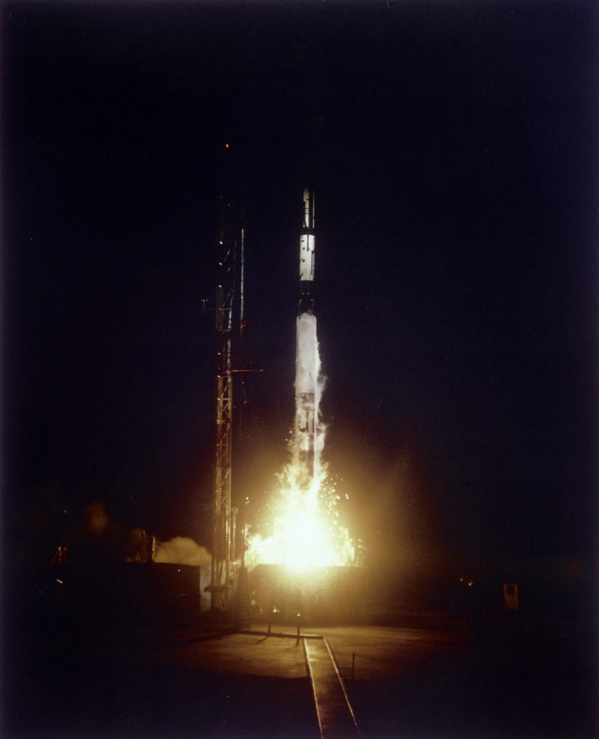 A slender three-stage Vanguard rocket lifts off at night beside its service tower.