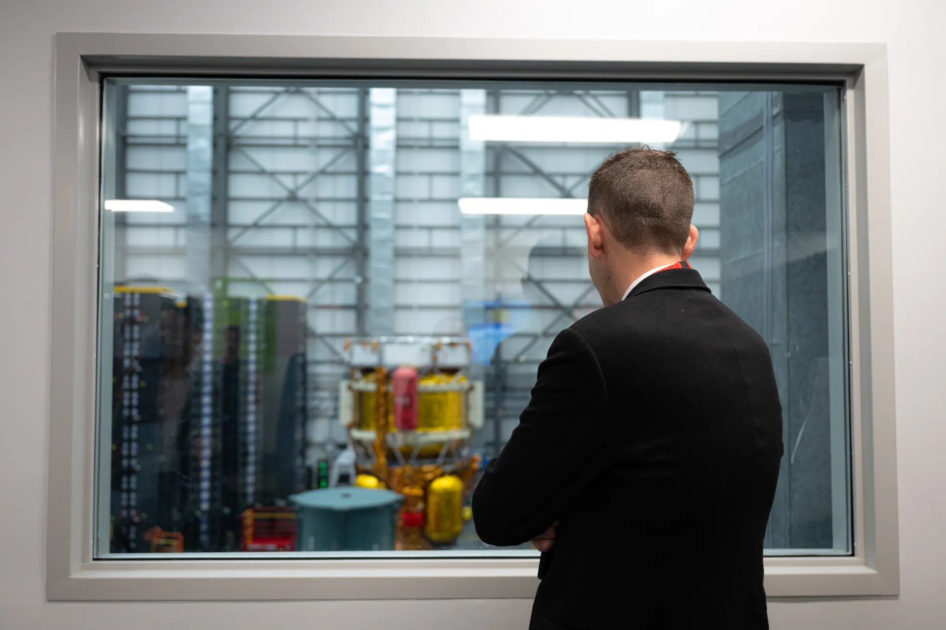 A man in a dark suit, seen from behind, looks through a large window at a gold-wrapped lunar lander in a clean room.