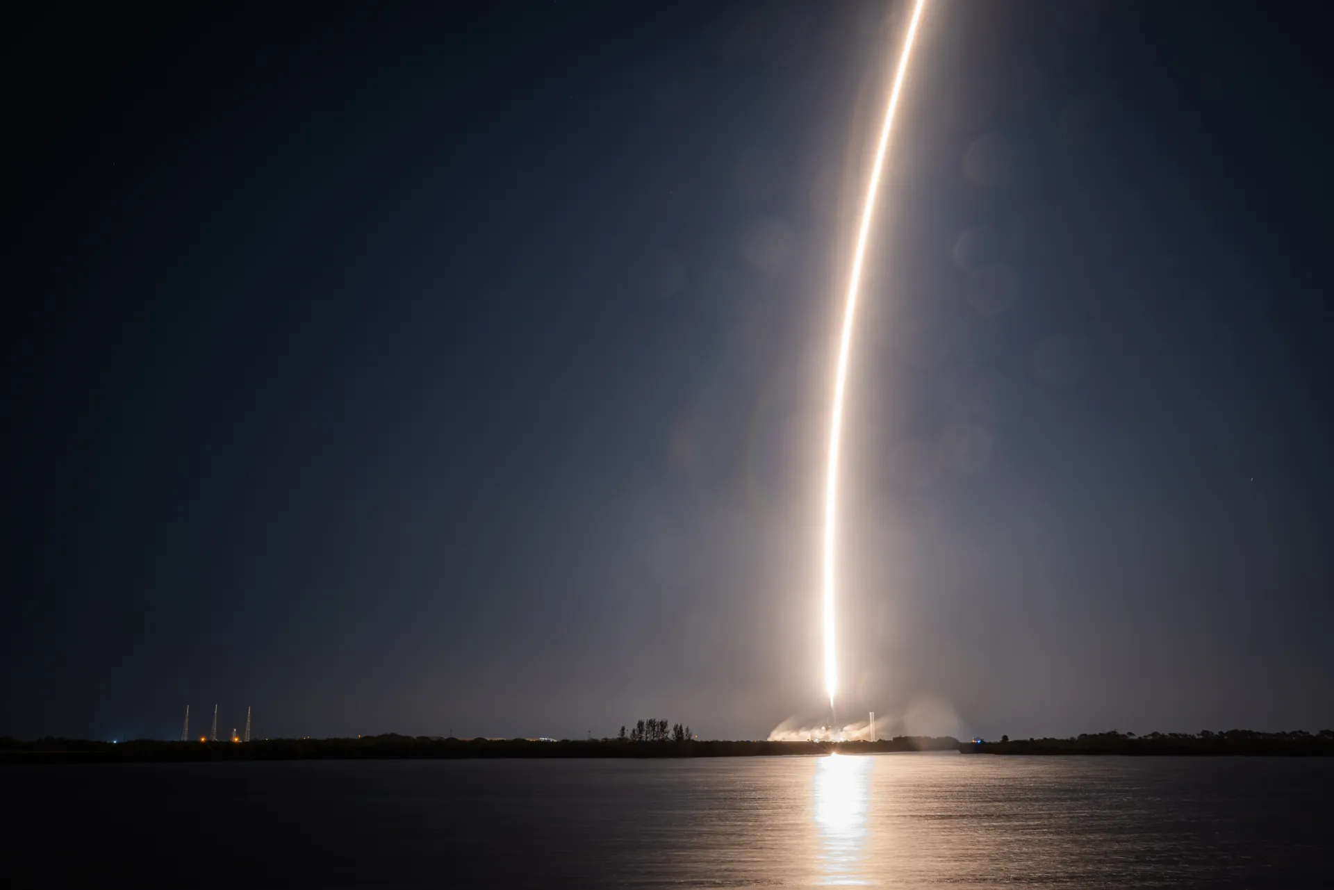 A long golden arc of rocket exhaust curves up from the horizon into a dark blue night sky over water.