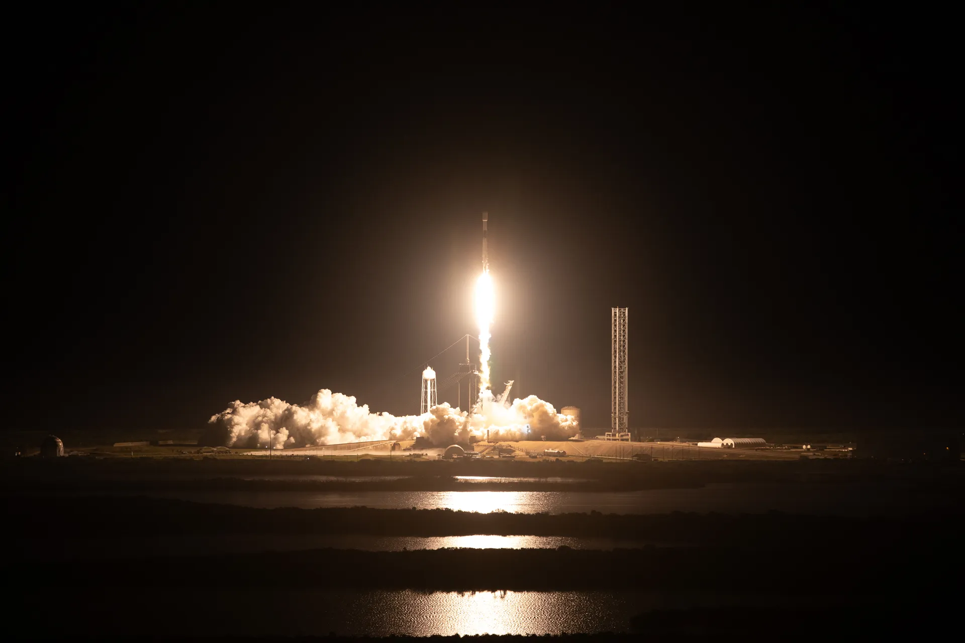 A Falcon 9 rocket rises at night from a coastal launch pad, its flame reflected in the water.