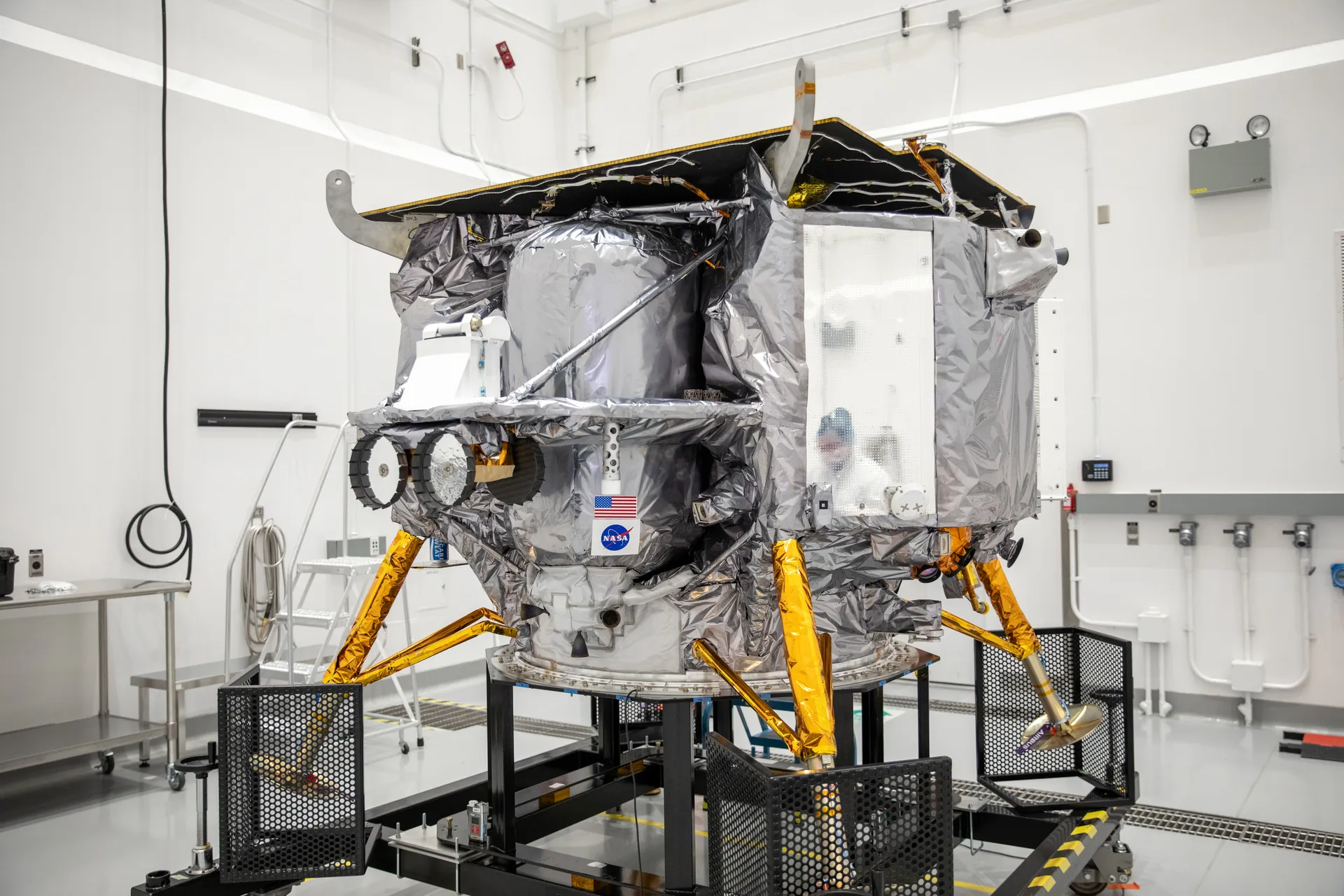Technicians in white clean-room suits work around a silver, boxy lunar lander on gold landing legs with a NASA logo on its side.