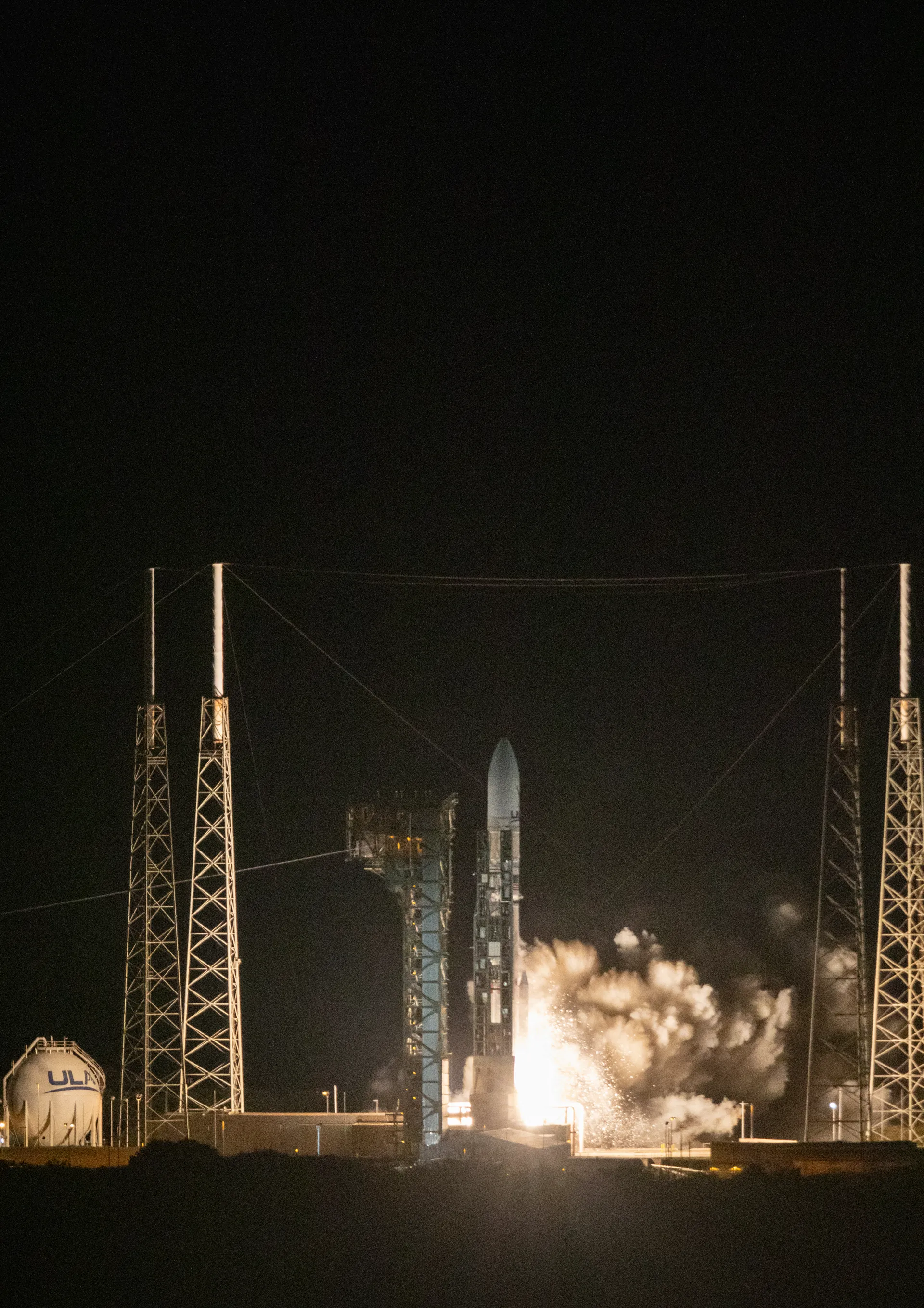 A rocket lifts off at night from a pad between four tall lightning towers, lit by its exhaust.