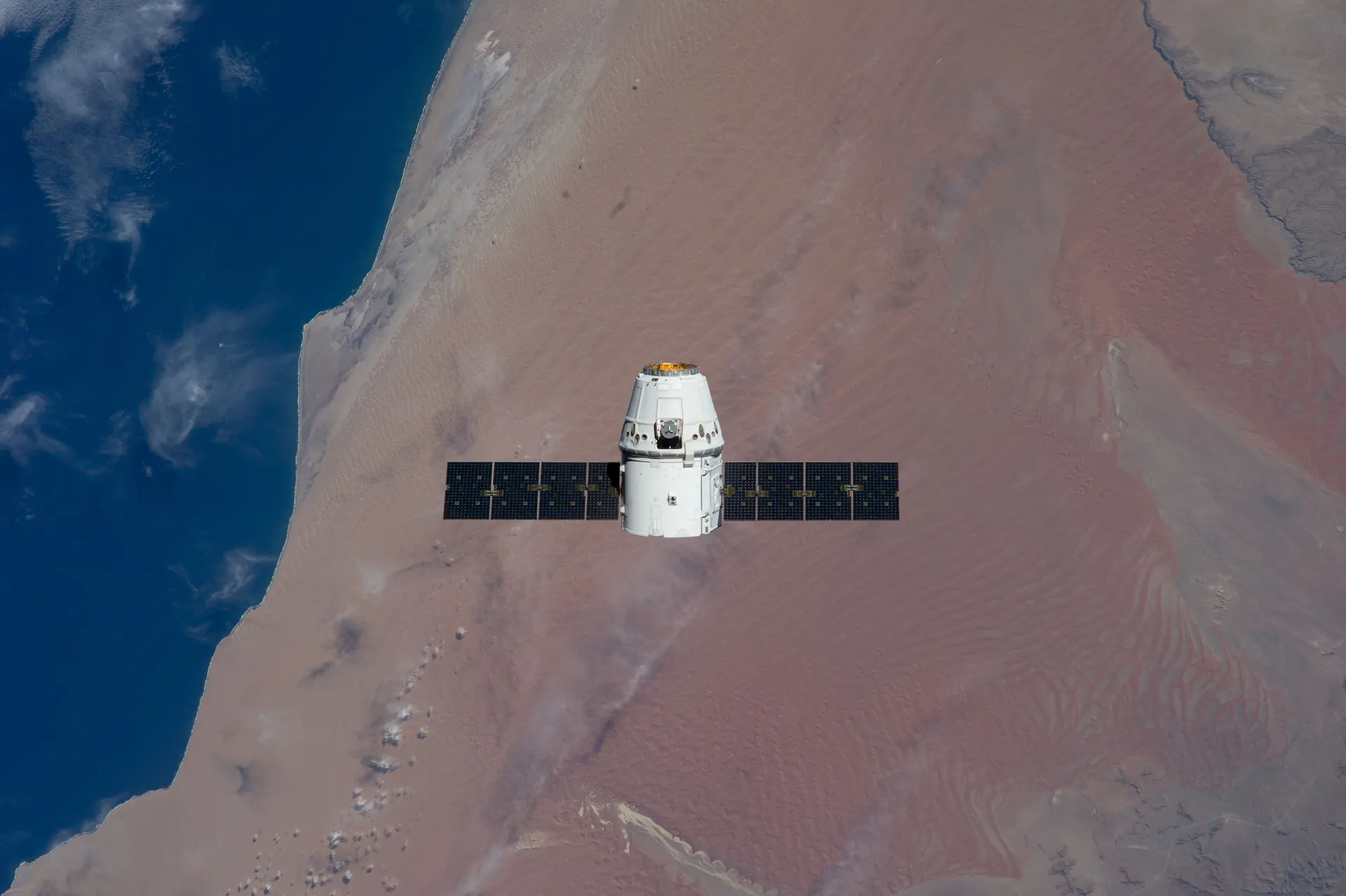 A white SpaceX Dragon capsule with two dark solar wings flies above the reddish Namib Desert and the blue Atlantic coast.