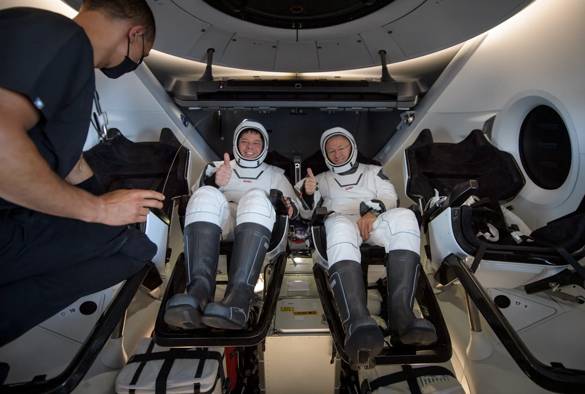Two astronauts in white pressure suits give thumbs-up from their seats inside a capsule while a technician kneels beside them.