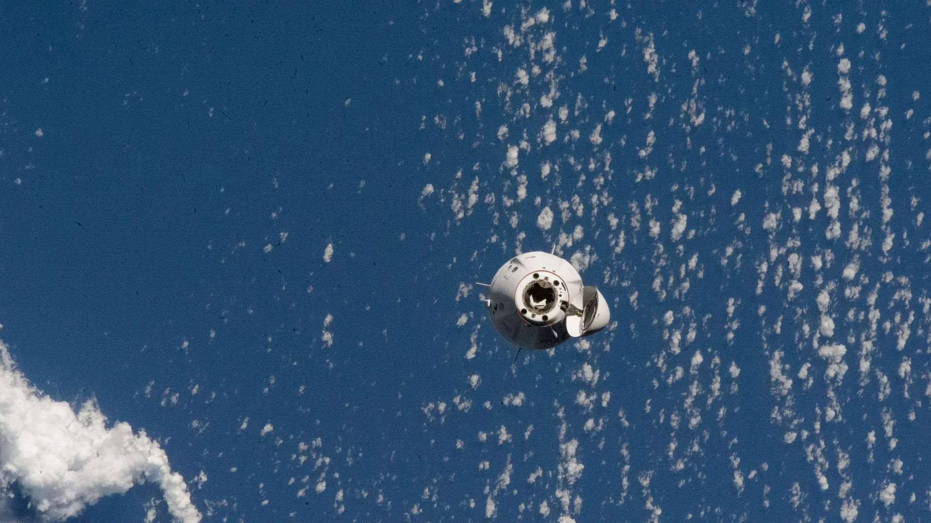 A white Crew Dragon capsule with its nose cone open approaches against a deep blue ocean dotted with small clouds.