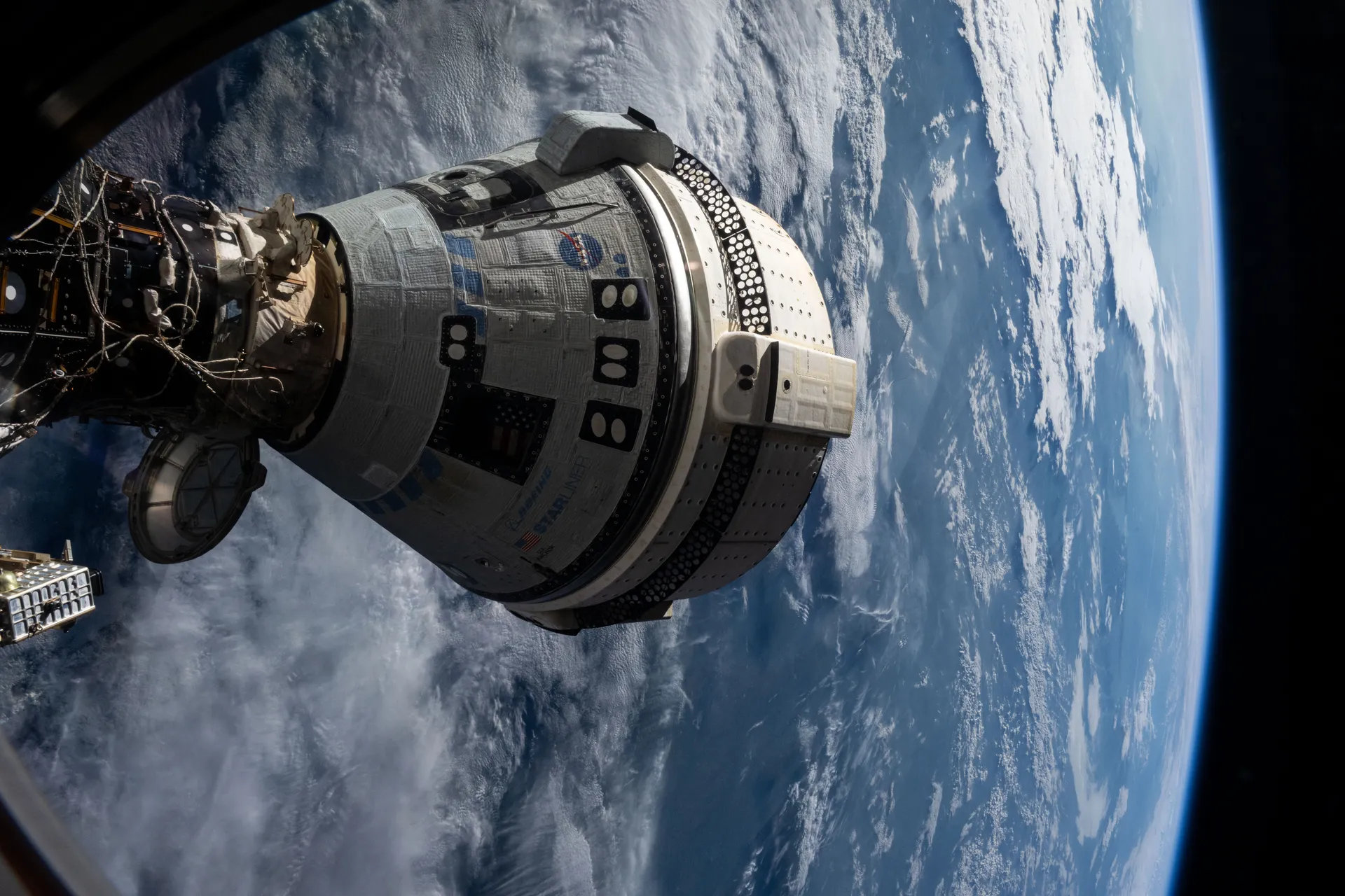 Boeing’s Starliner capsule, white with dark panels, docked to the station high above a blue and white Earth, framed by part of the station.