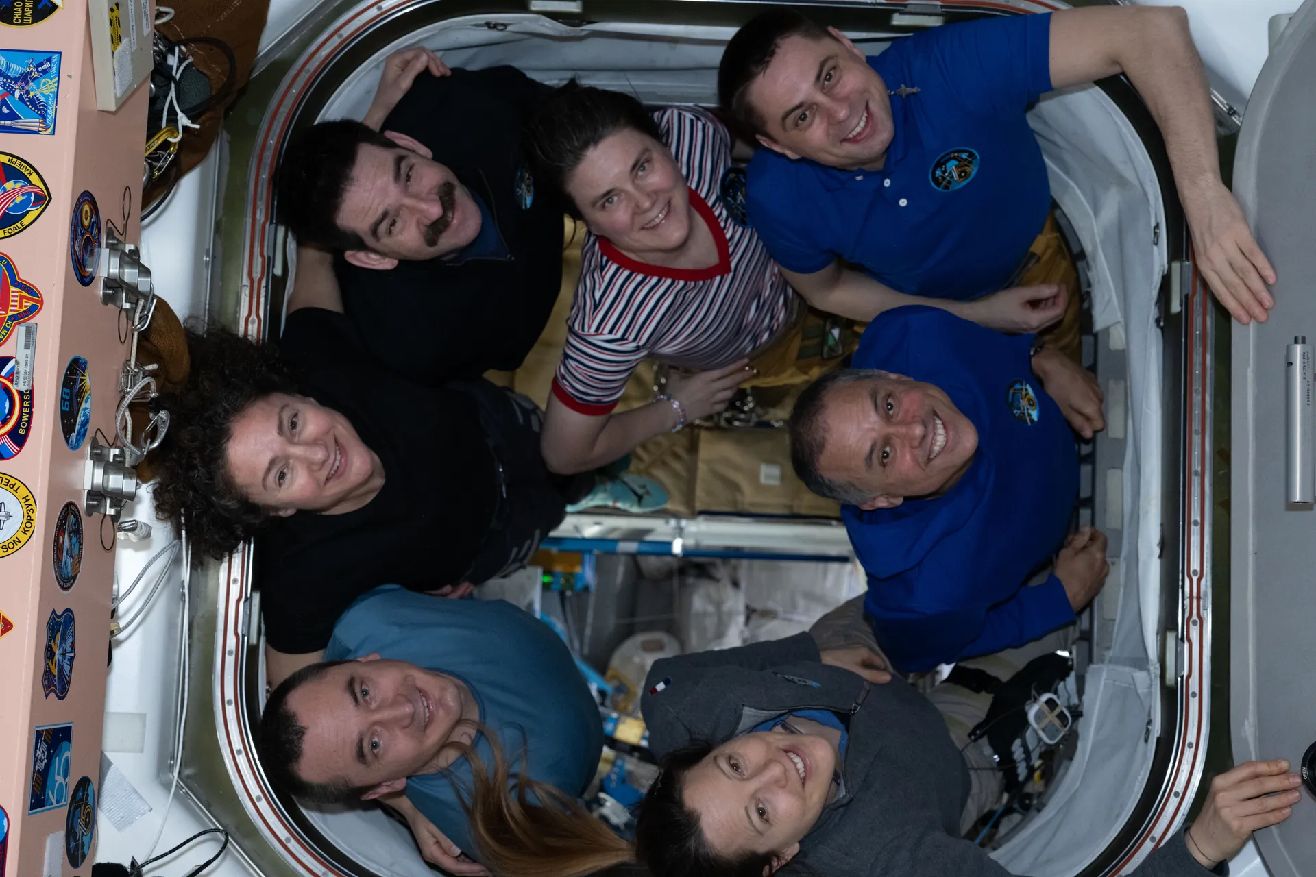 Seven crew members in shirts of different colors crowd together in a round hatchway, smiling at the camera.