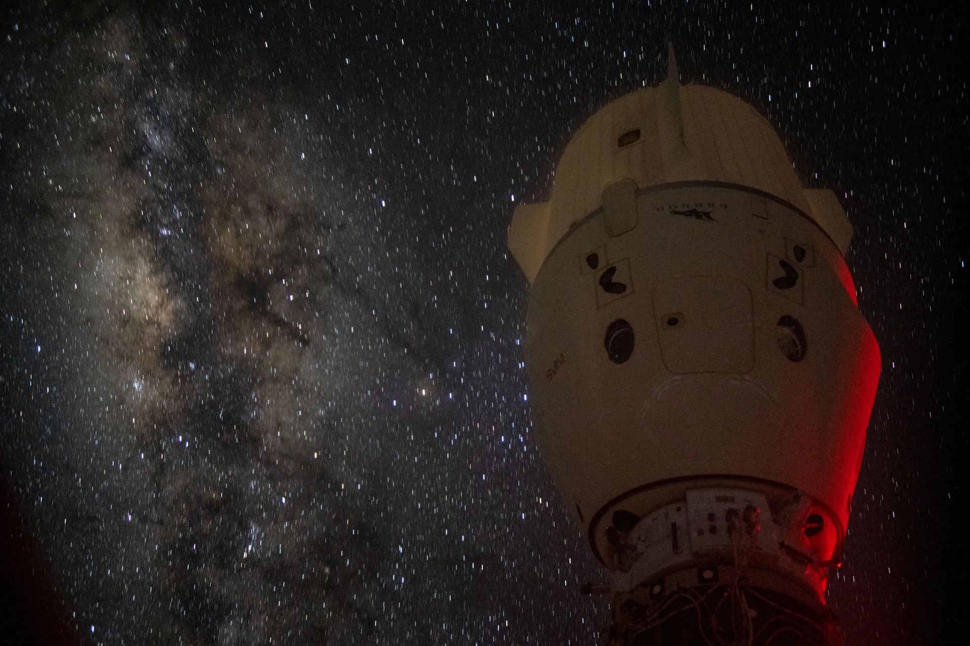A SpaceX Crew Dragon docked to the space station, dimly lit, with the Milky Way behind it.