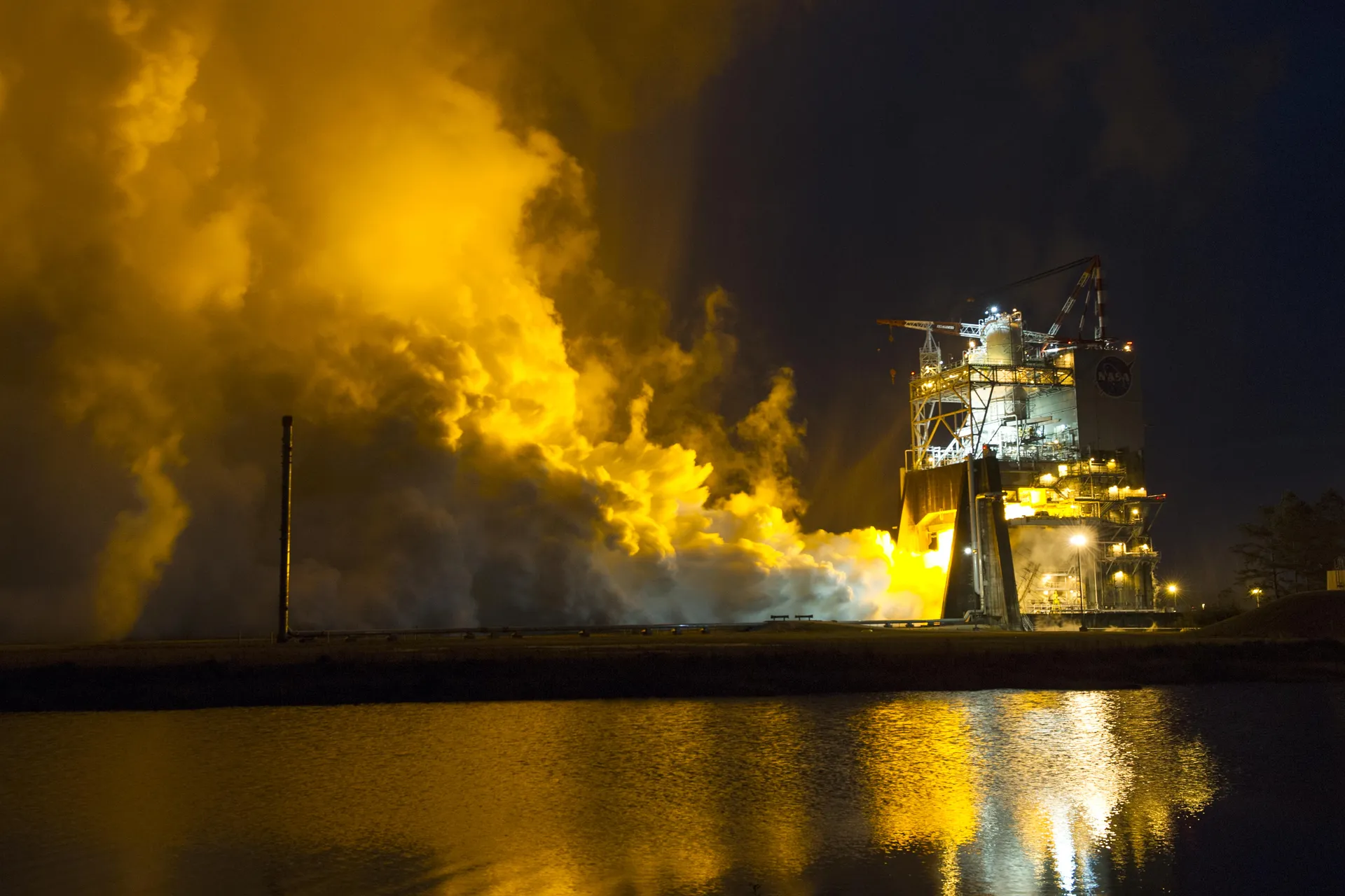 An RS-25 engine fires on a test stand at night, lighting a vast cloud of steam yellow and reflecting in the water below.