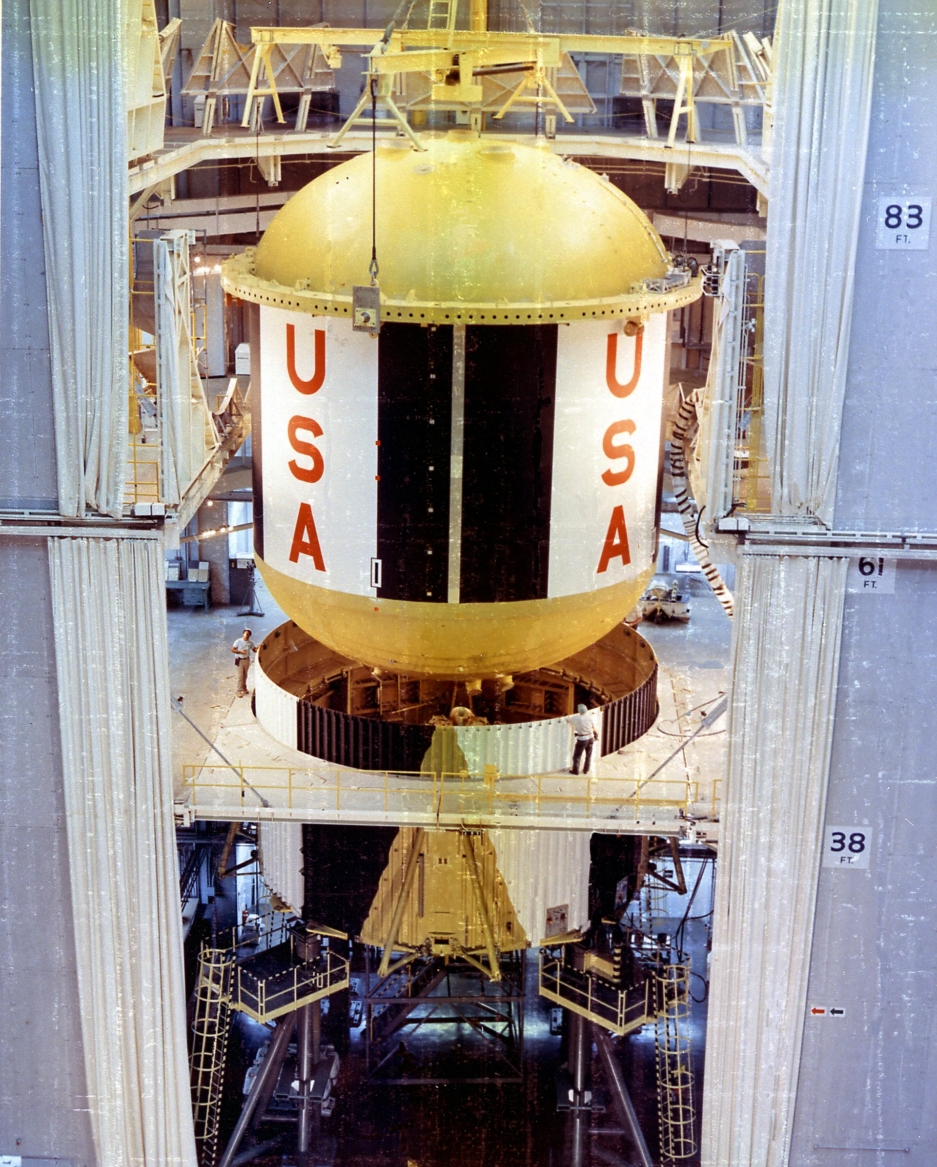 A huge Saturn V first-stage fuel tank, marked USA, is lowered into place inside a tall assembly building.