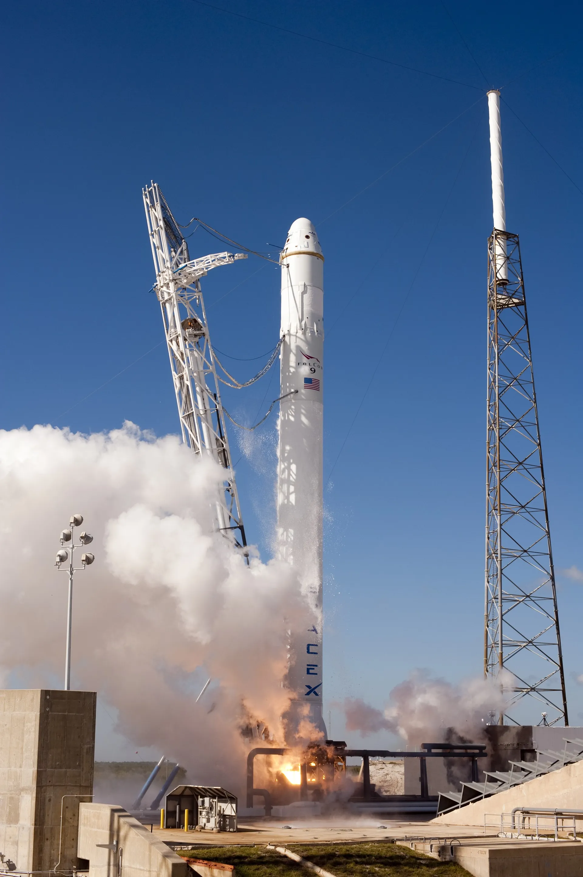 White Falcon 9 on its pad at Space Launch Complex 40 with a cloud of exhaust billowing from its base during an engine test, blue sky behind.