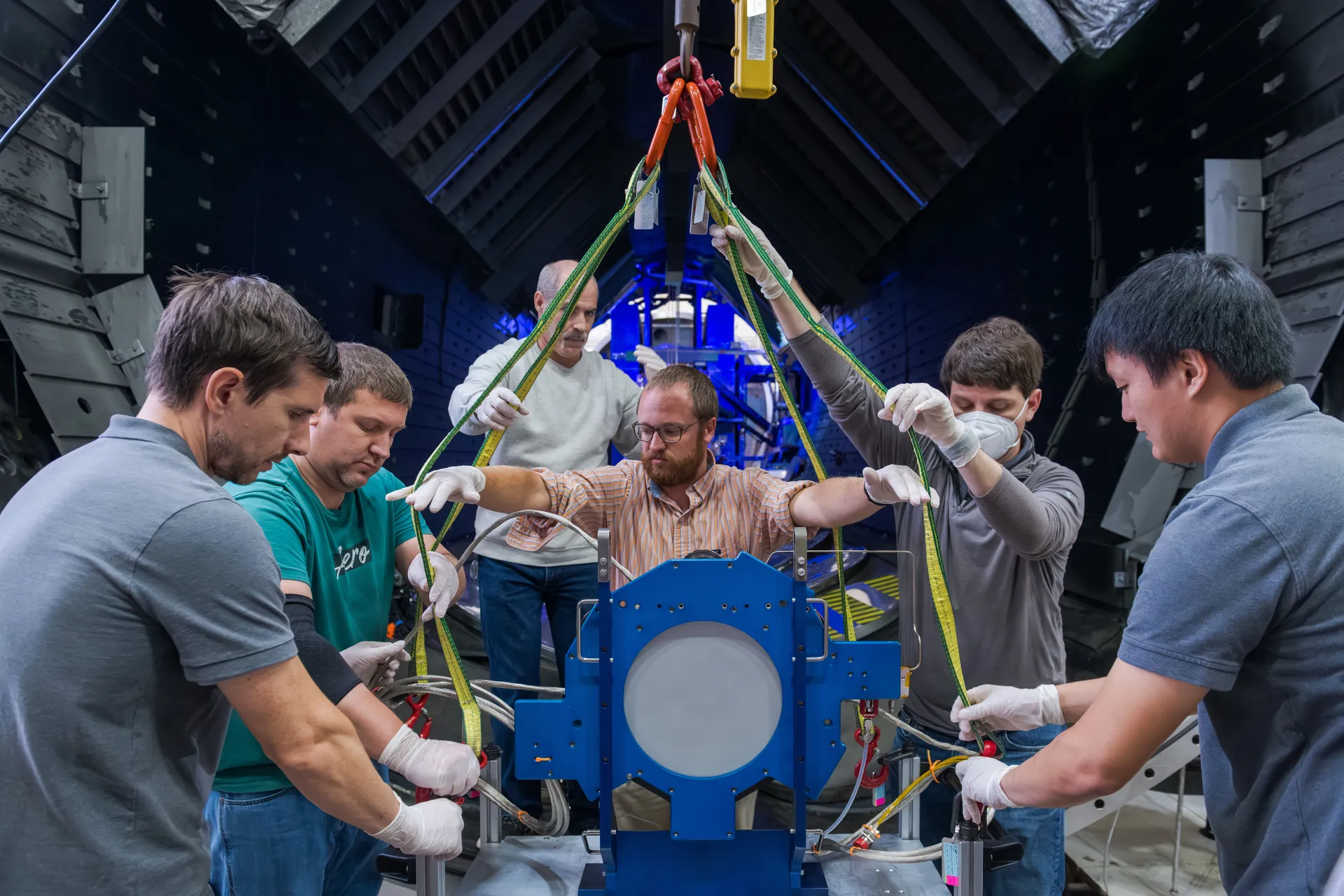 Six engineers in a dark test chamber guide straps holding a thruster in a blue mounting fixture, with blue light behind them.