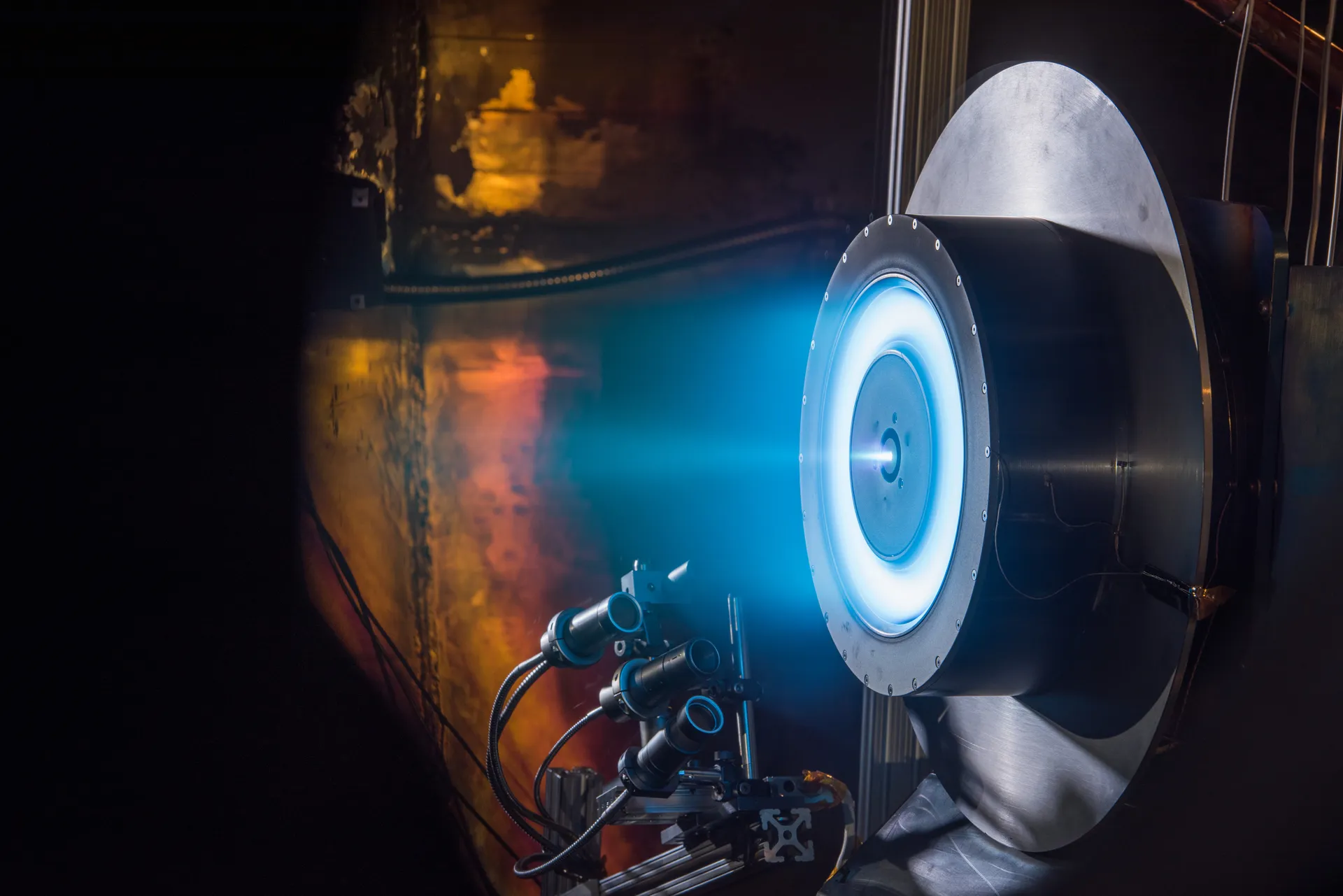 A large round Hall thruster glows with a blue-violet plasma plume inside a dark vacuum chamber, with cameras on a mount in the foreground.
