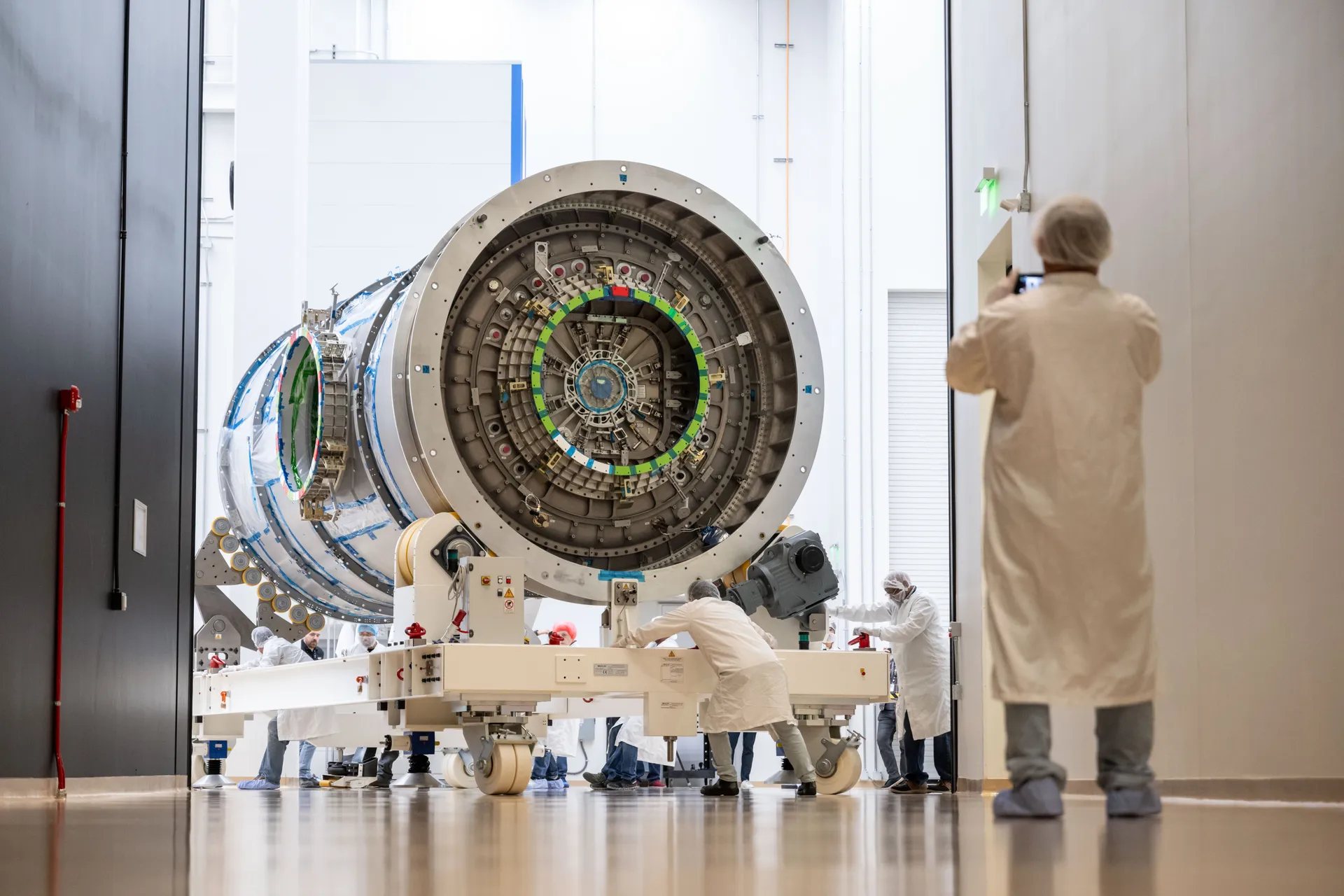 Technicians in white cleanroom suits push HALO, a large silver cylinder with a docking port facing the camera, on a wheeled frame through a high doorway.
