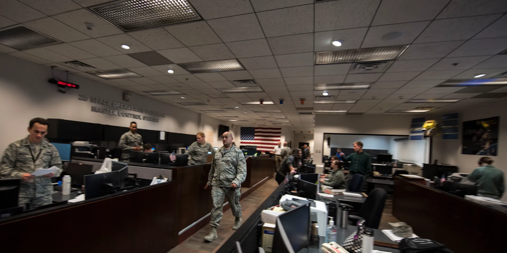 A long operations room with rows of consoles; airmen in uniform work at screens beneath a wall sign reading ‘2D Space Operations Squadron Master Control Station’, with a U.S. flag at the far end.