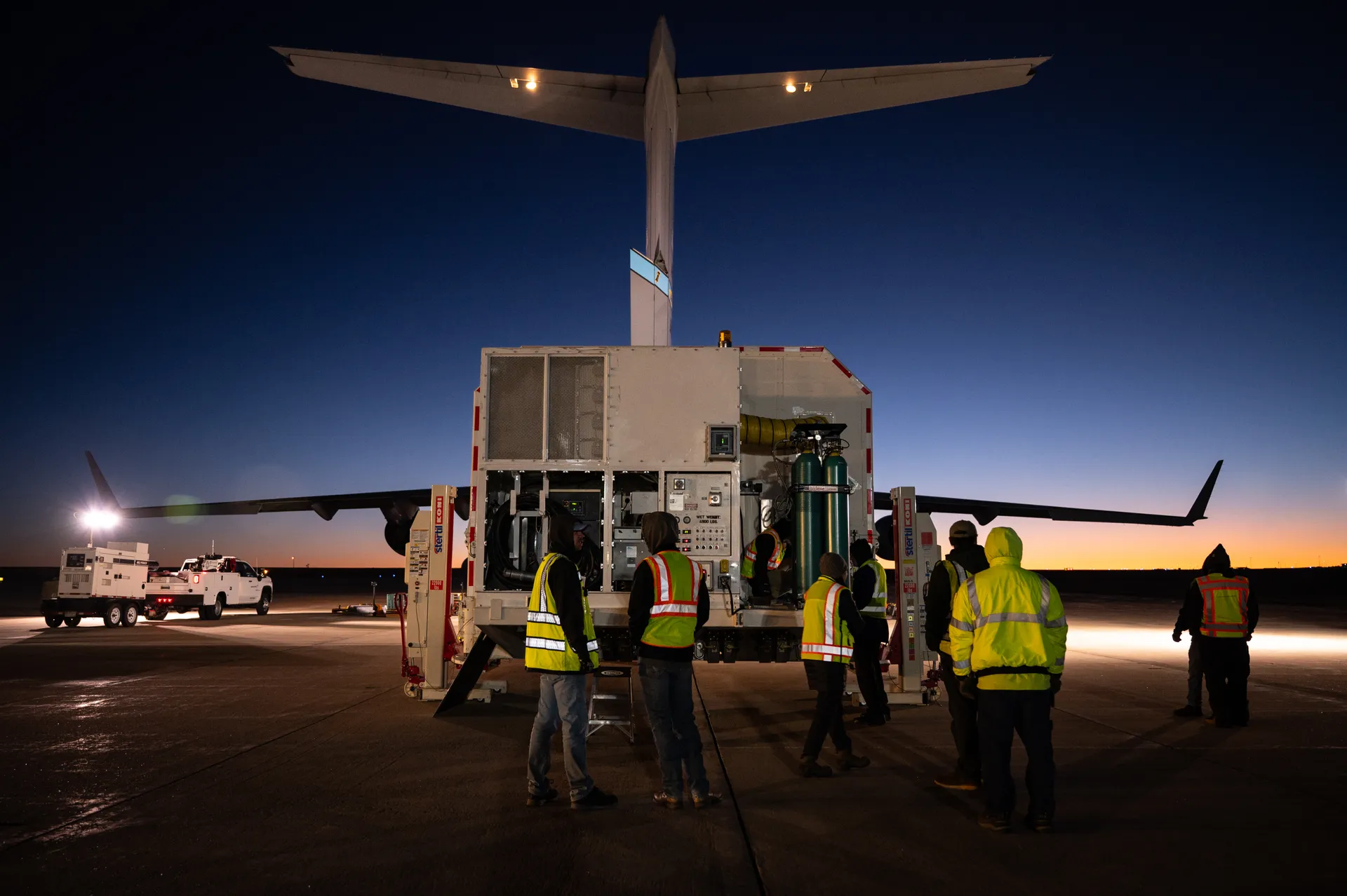 At dusk on an airfield, workers in reflective vests stand around a large white shipping container on a loader behind the open tail of a C-17 cargo plane.
