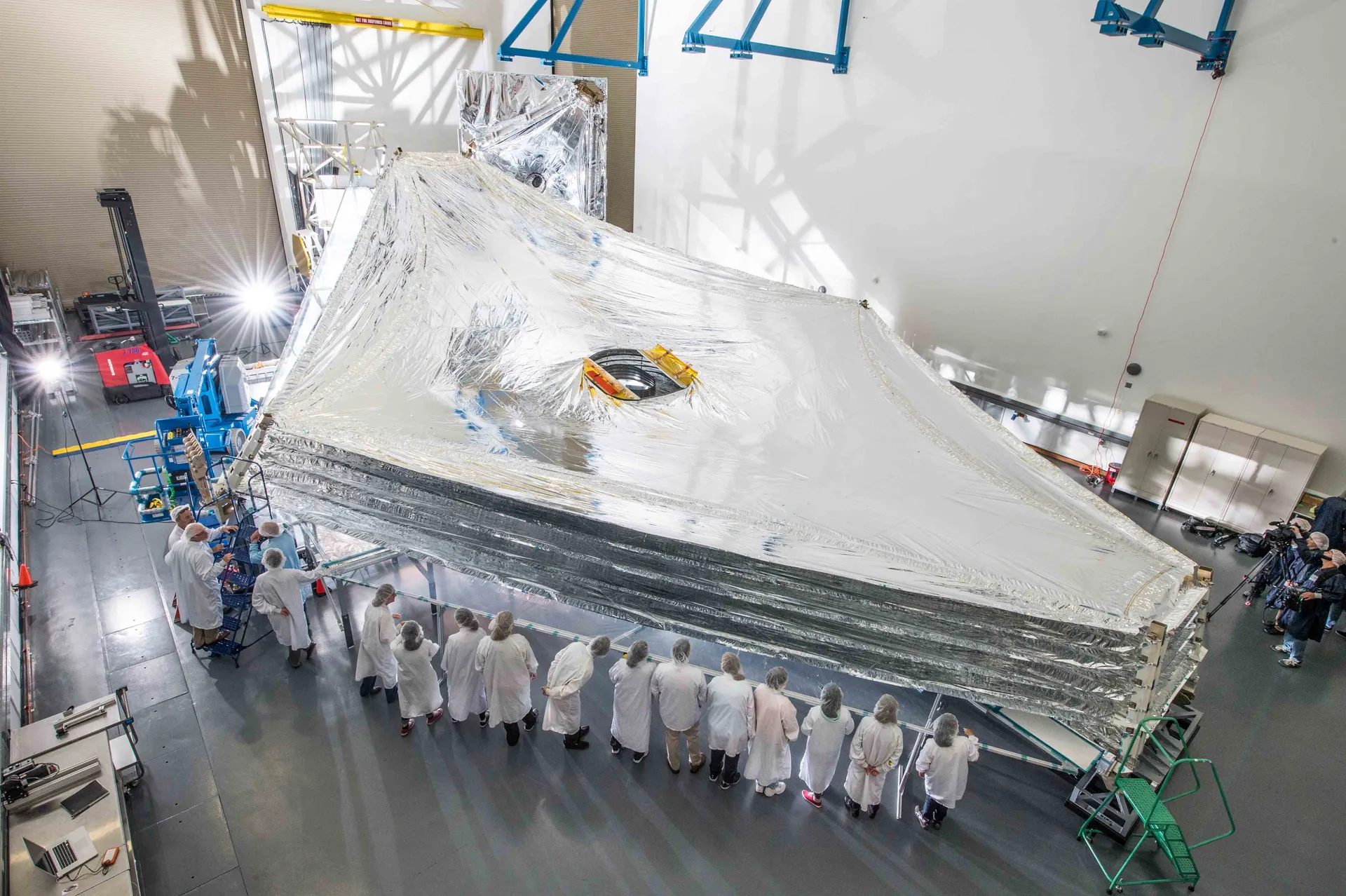 A wide, silvery, five-layer membrane is stretched out across a large clean room while a long row of technicians in white suits stands along its edge.