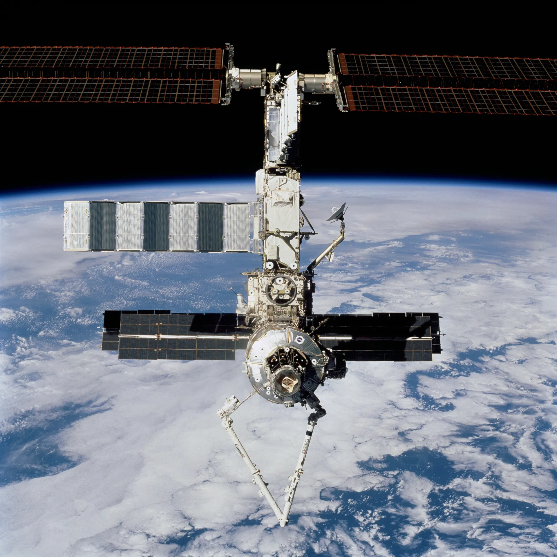 The station seen from below against a white and blue Earth, with the new robotic arm extended downward.