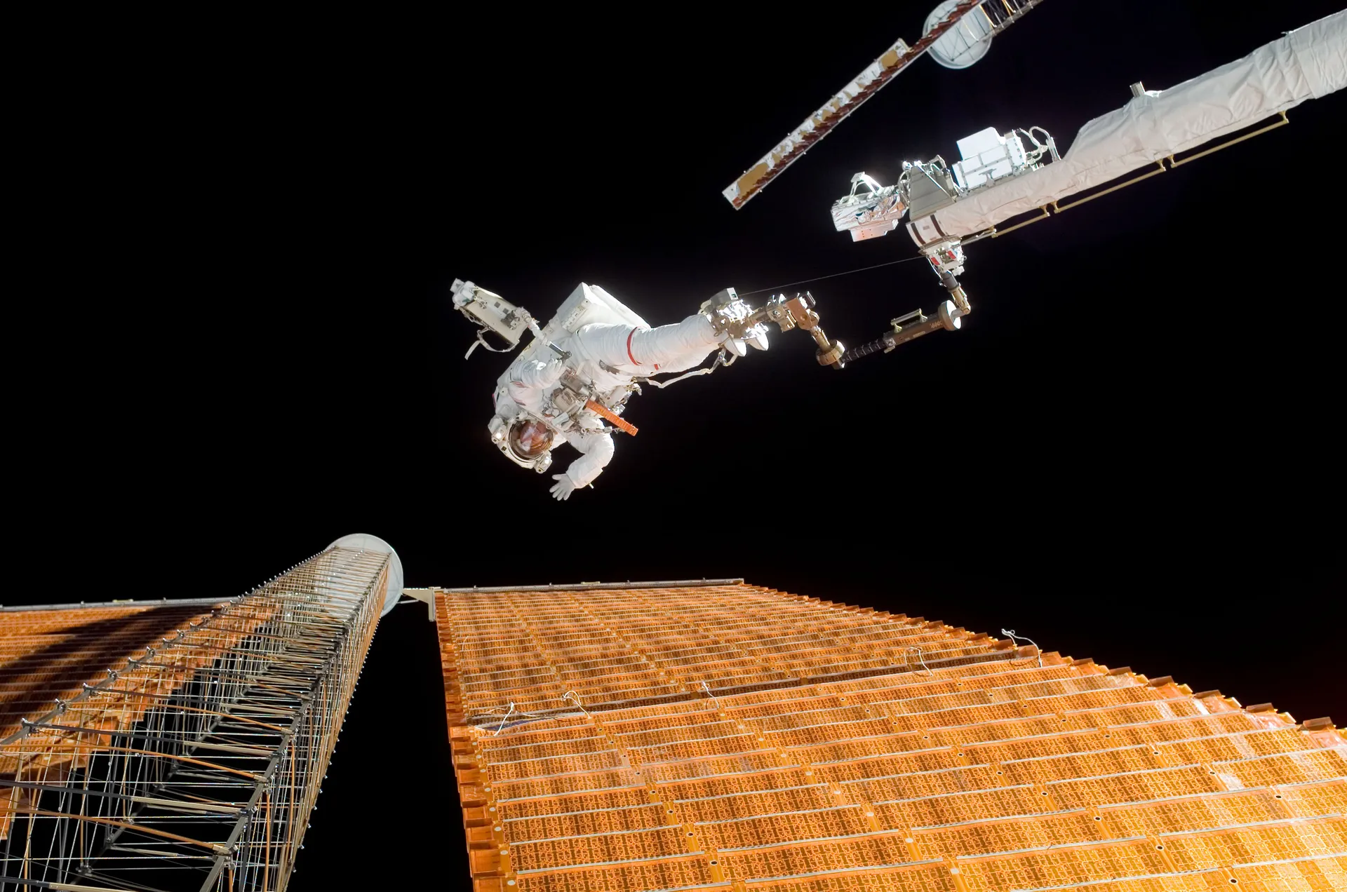 An astronaut in a white spacesuit stands at the end of a long boom beside a vast orange solar array, against black space.