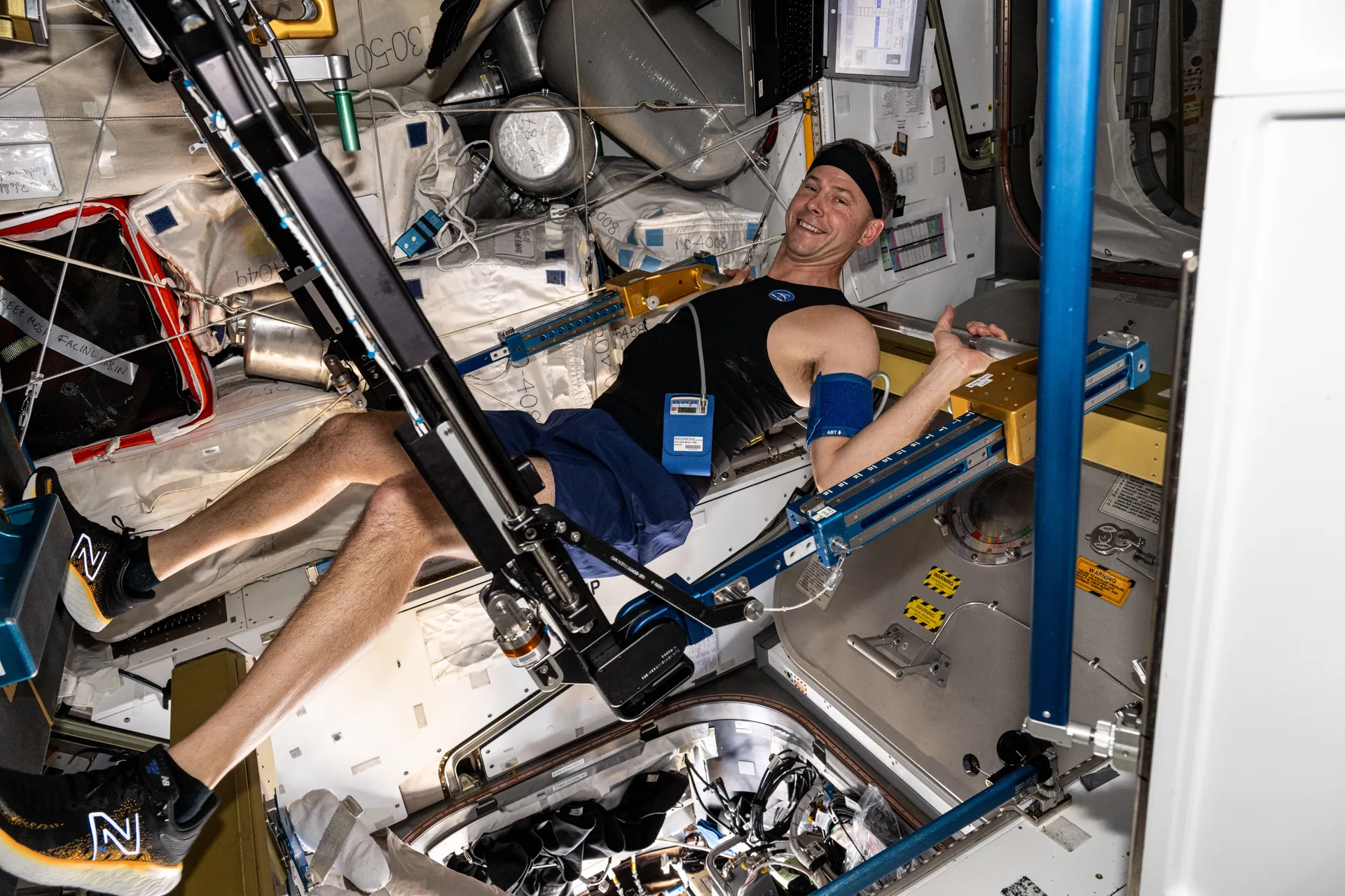 Nick Hague lies under the bar of the Advanced Resistive Exercise Device, legs braced, wearing a sensor headband.