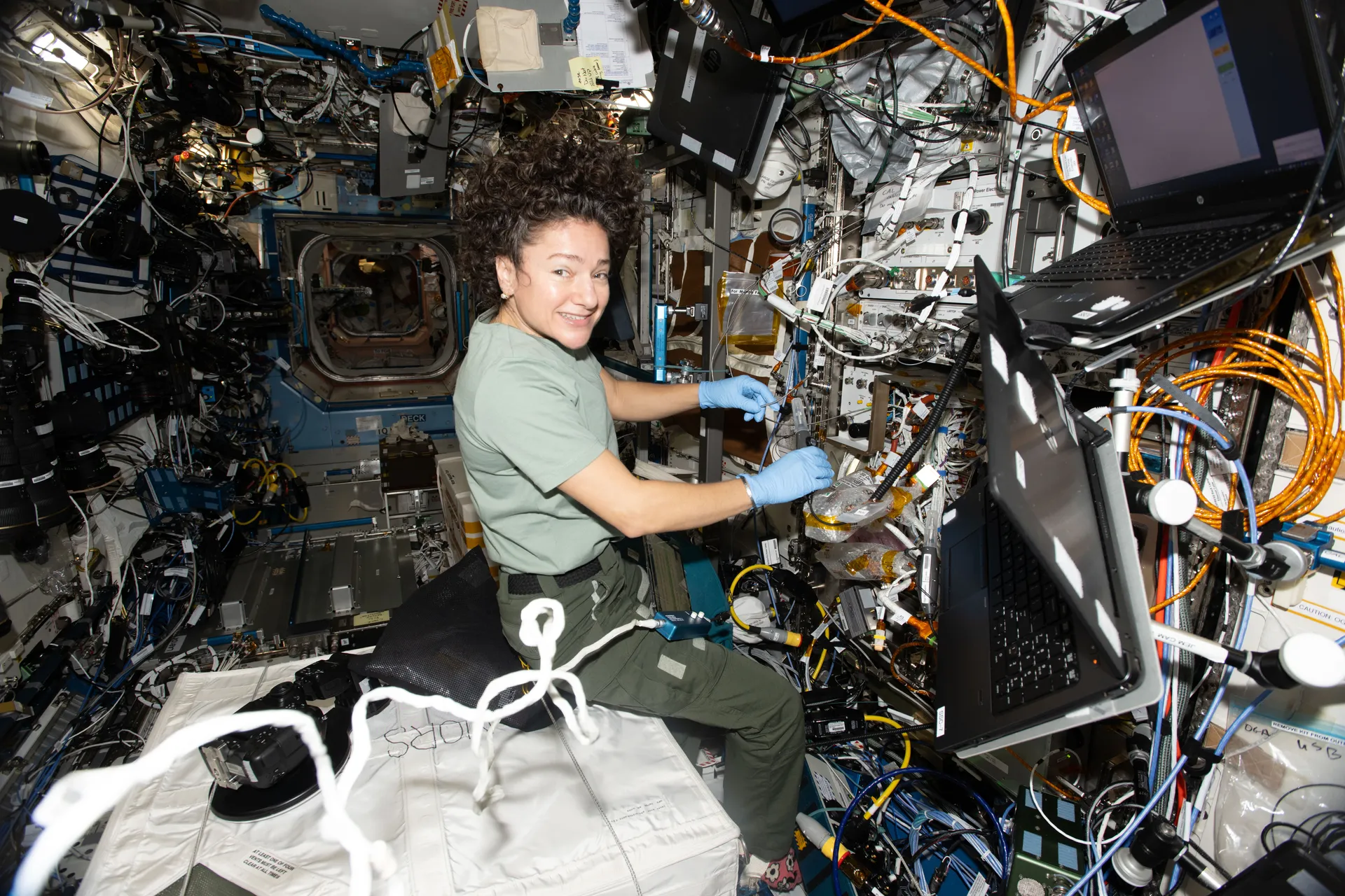 Jessica Meir, wearing blue gloves, works inside a rack crowded with orange fiber cables and electronics.