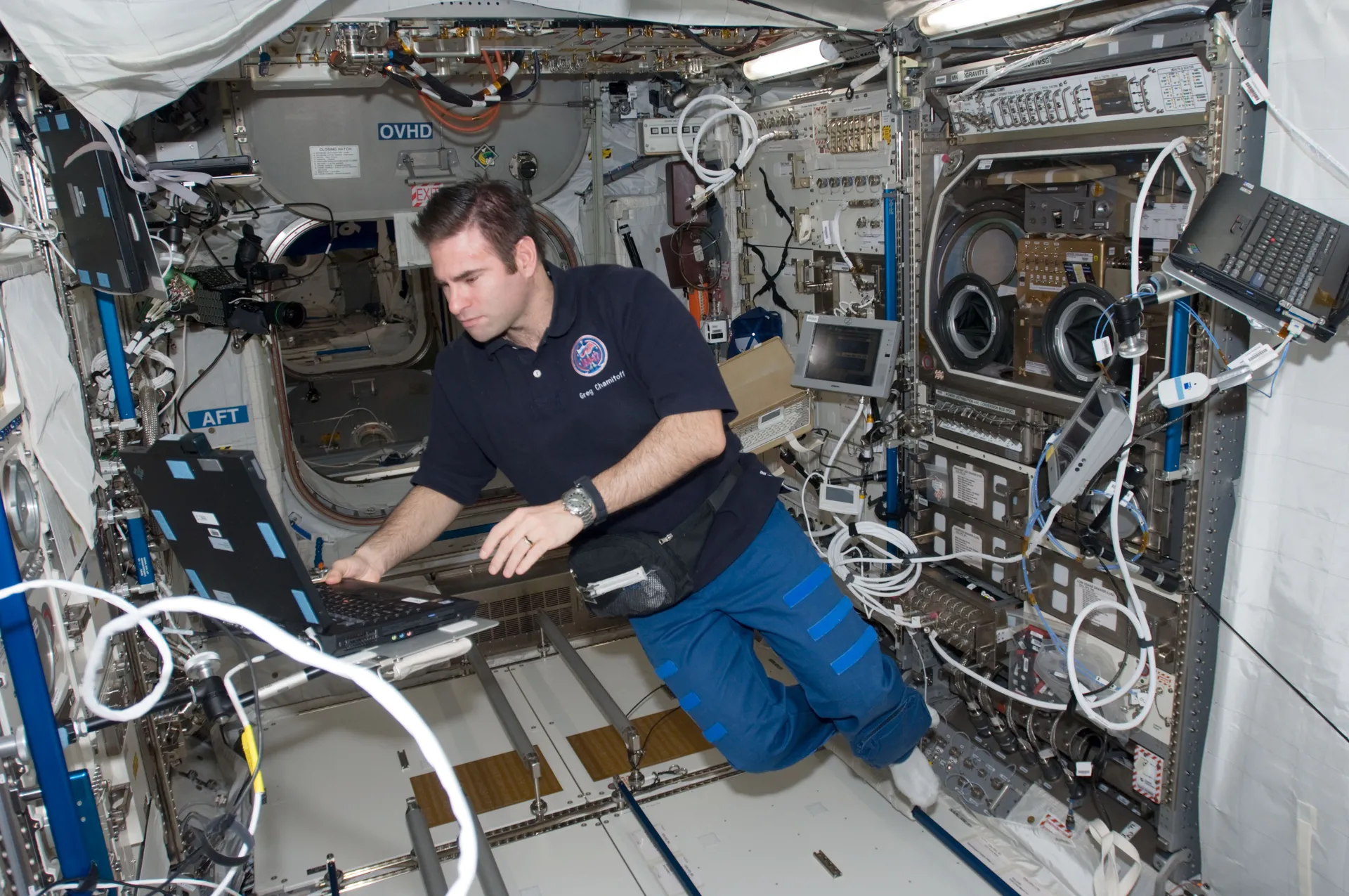 Greg Chamitoff types on a laptop beside a large rack whose front panel has round glove ports.
