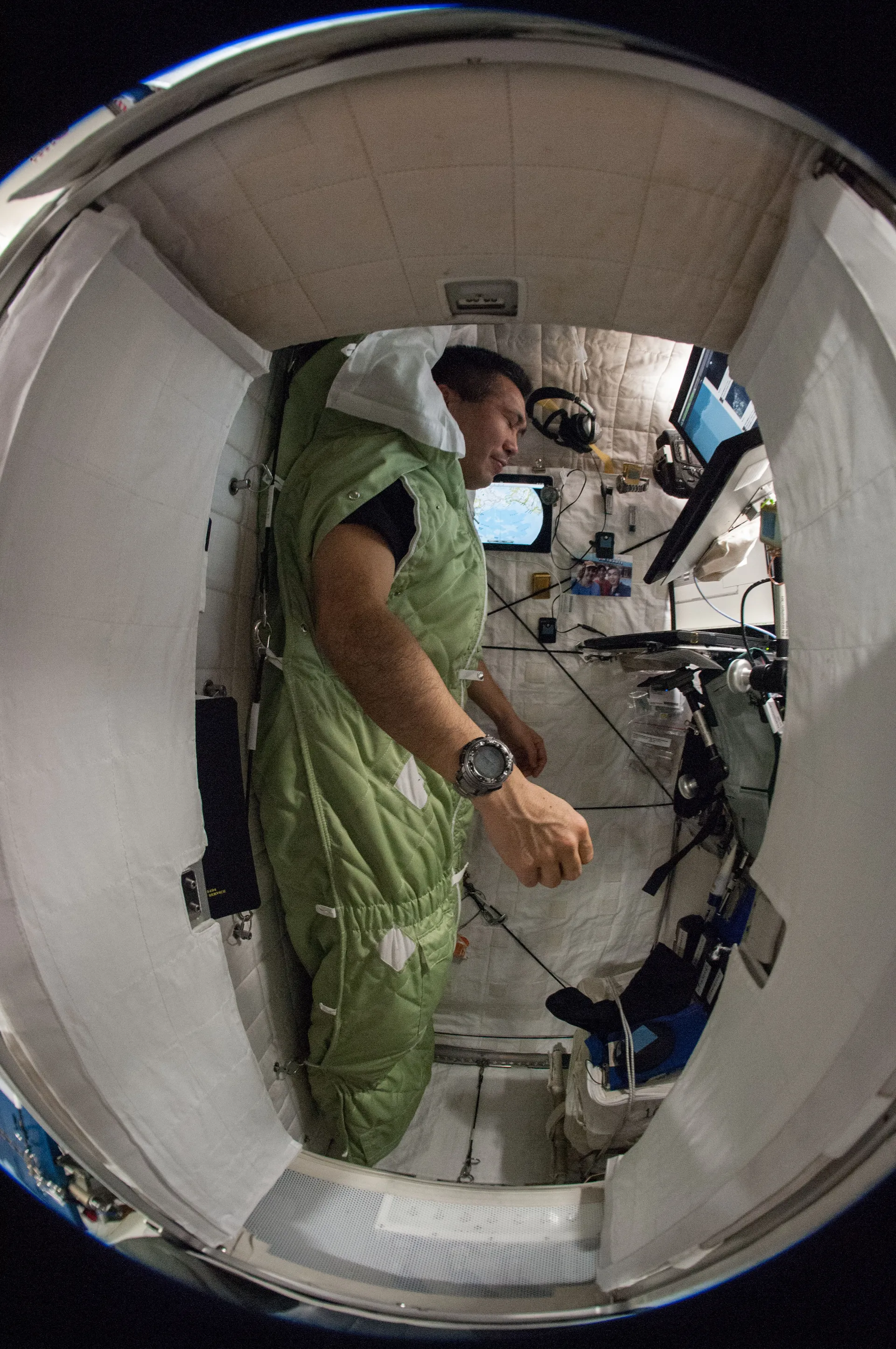Koichi Wakata, zipped into a green sleeping bag fastened to the wall of a small white sleep station, seen through a fisheye lens.