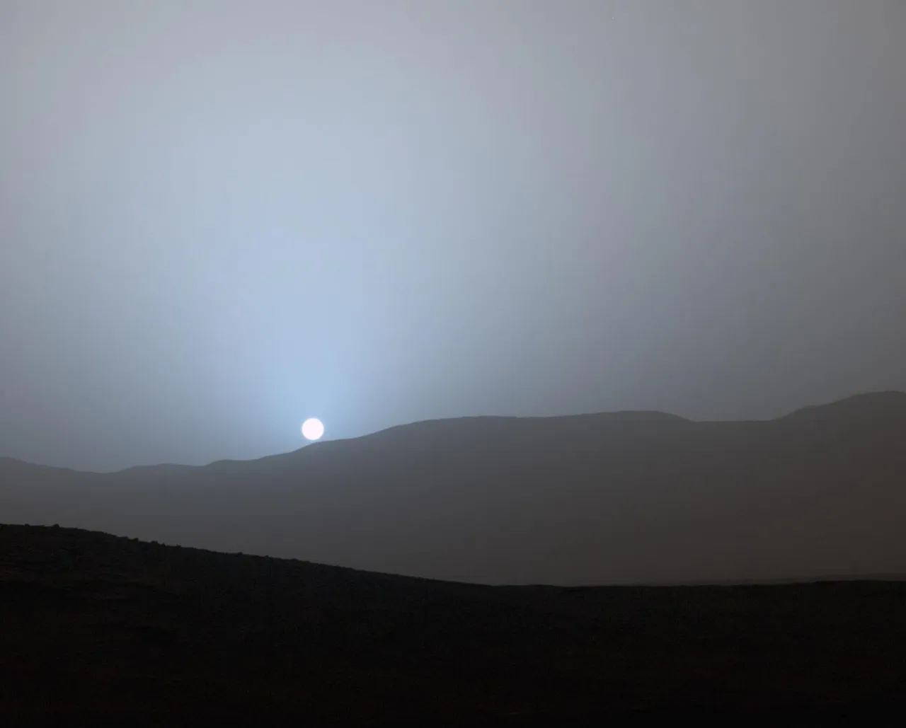 A small white Sun sets behind dark crater hills, surrounded by a blue glow in a gray sky.