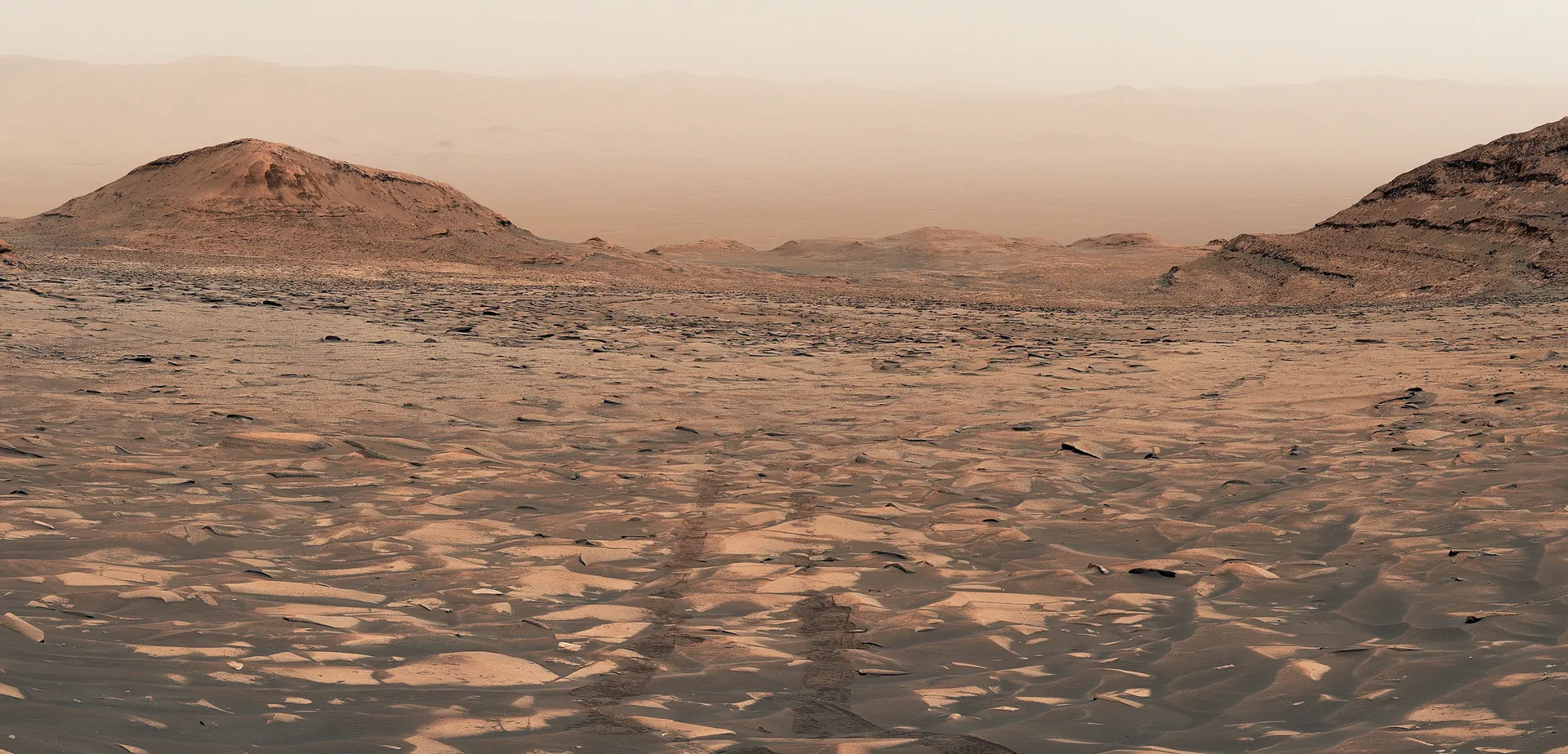 A wide view down a broad, sandy valley on Mars between dark hills, with rover tracks leading away toward a hazy crater floor and distant rim.