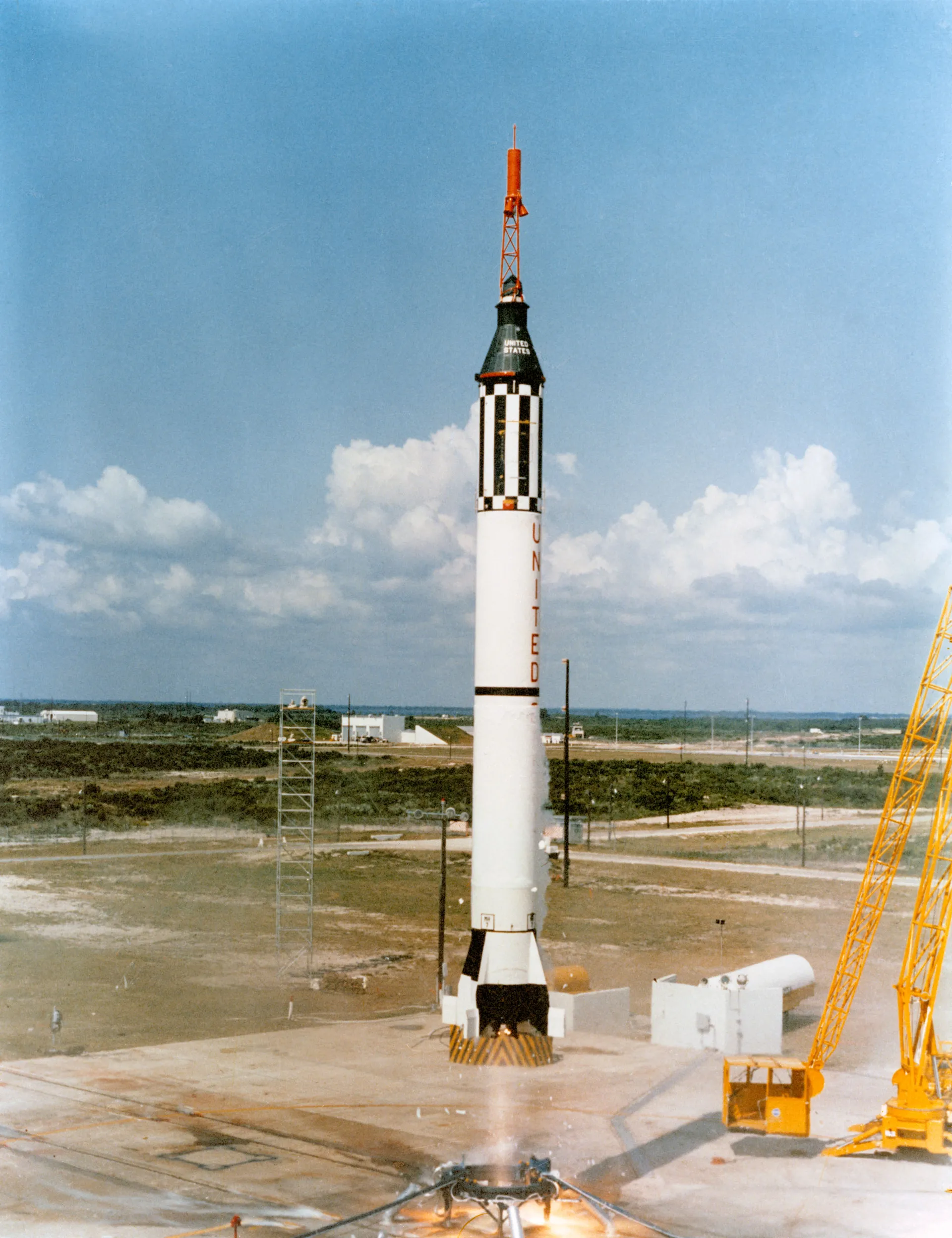 A slender black-and-white Mercury-Redstone rocket rises from its pad on a clear day.