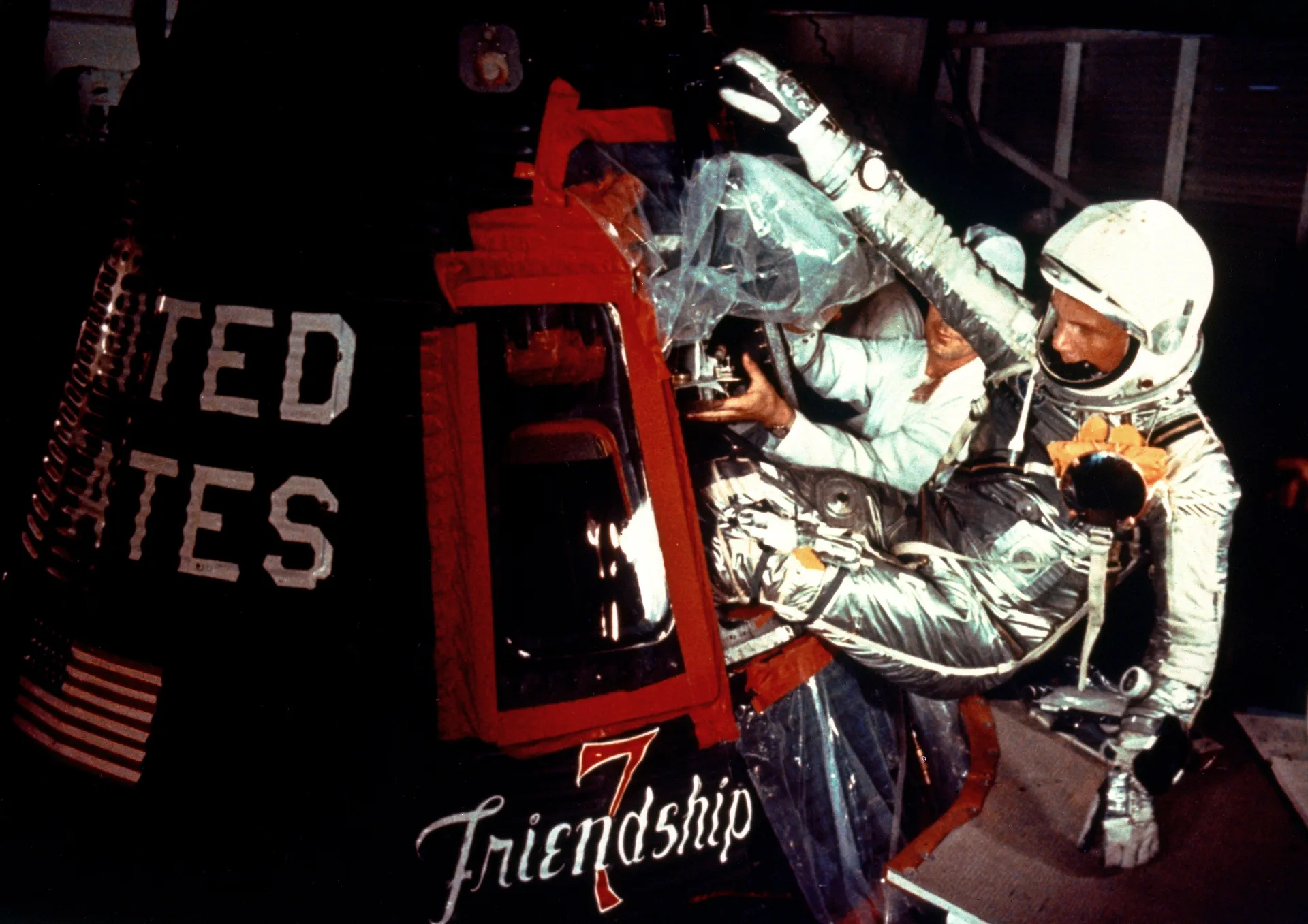 John Glenn in a silver pressure suit climbs into the hatch of Friendship 7, beside the painted capsule name.
