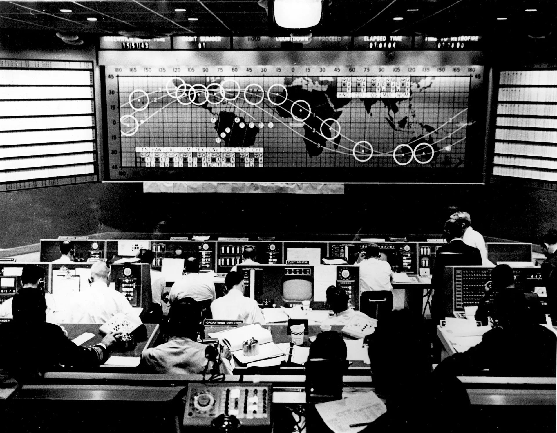 Flight controllers at rows of consoles in the Mercury Control Center, facing a wall map of the world with the capsule’s orbital track.