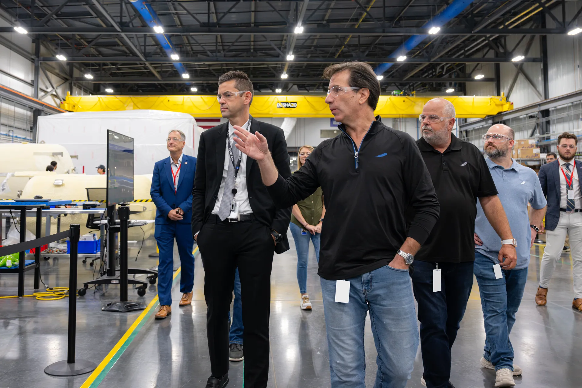 NASA’s administrator and Blue Origin managers walk through a bright factory hall with white spacecraft hardware behind them.