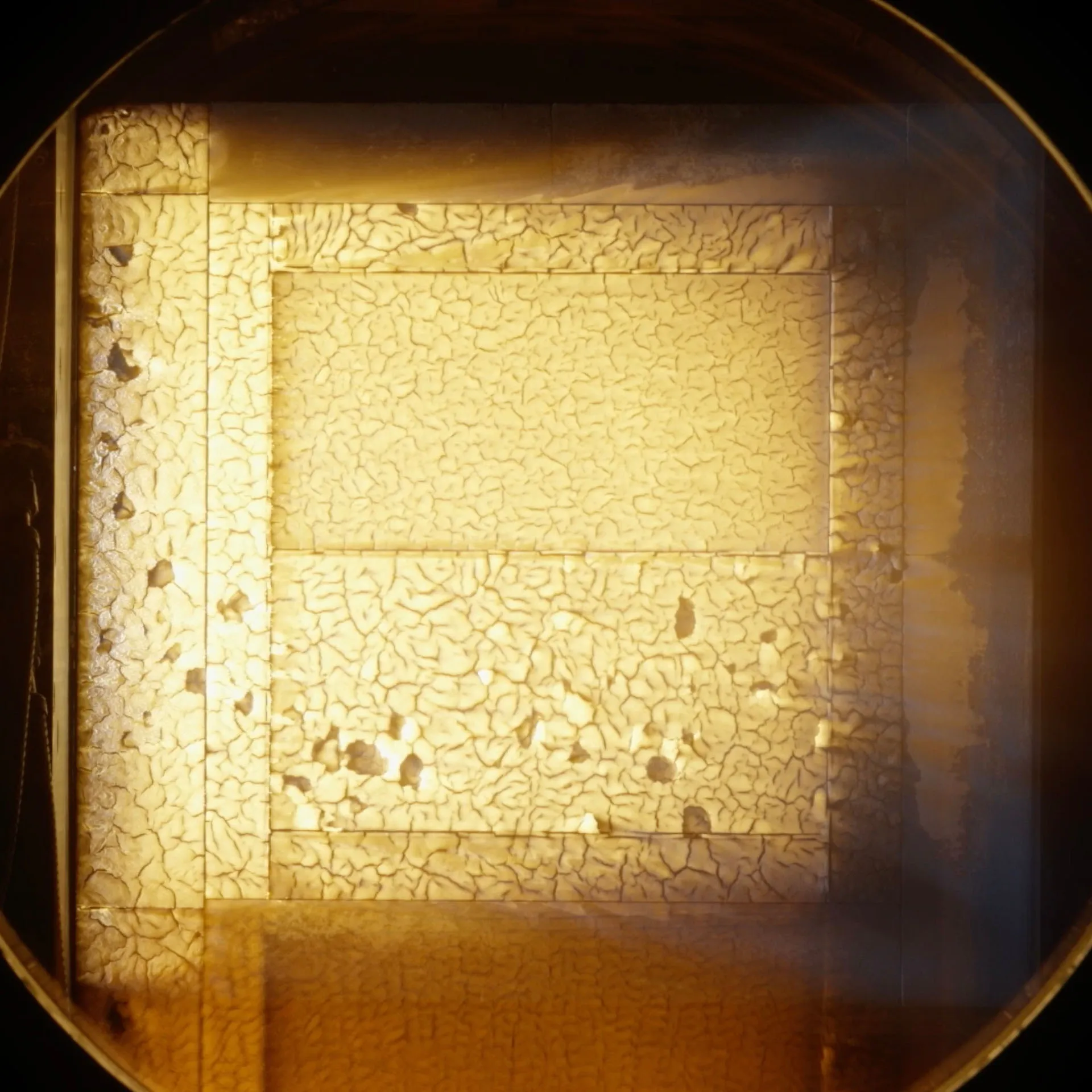 A square test block glows orange-white inside the round window of an arc jet chamber; the upper half has a smoother surface than the cracked lower half.