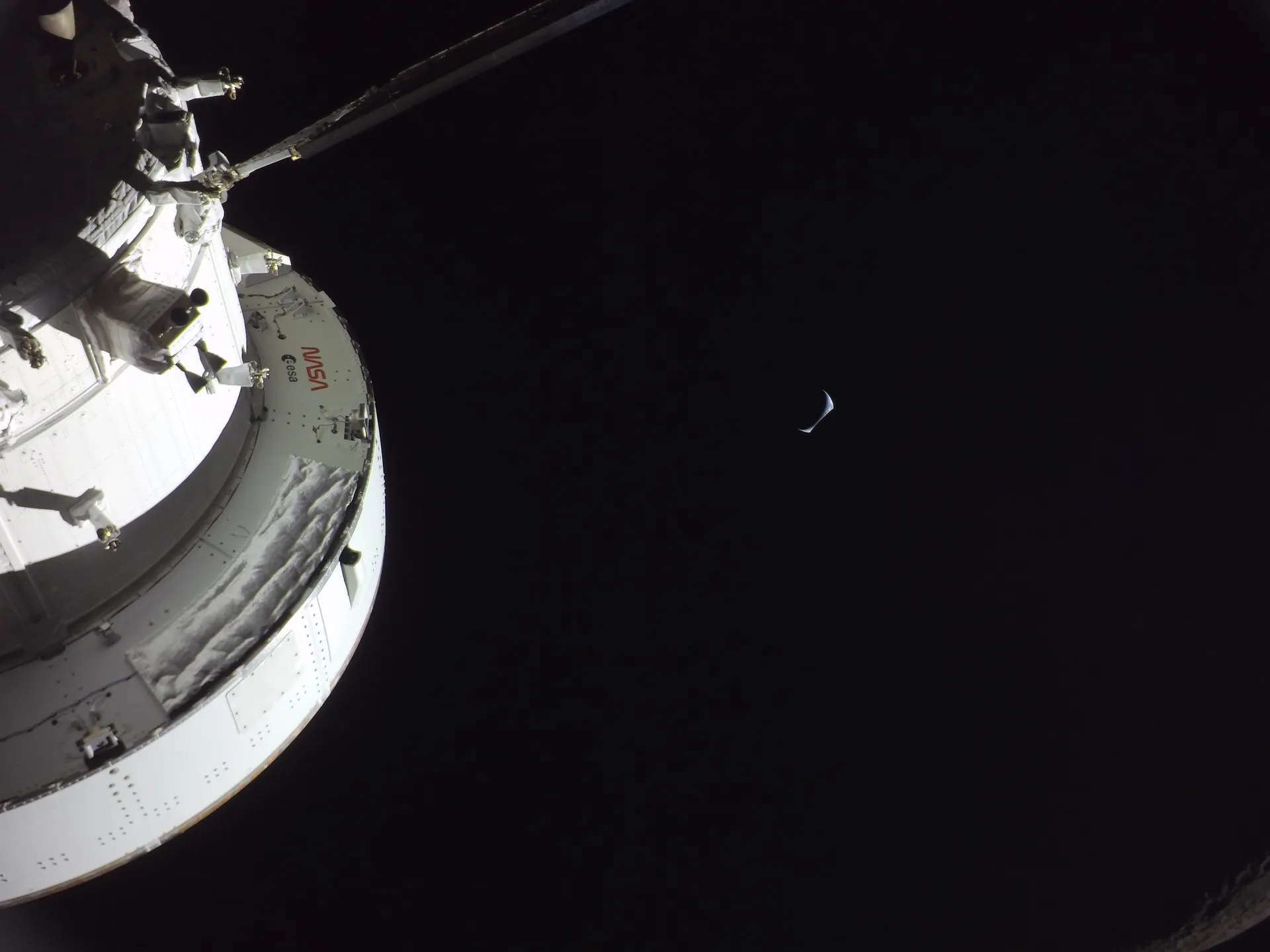 A camera on Orion’s solar array looks along the spacecraft toward a thin crescent Earth rising in black space.
