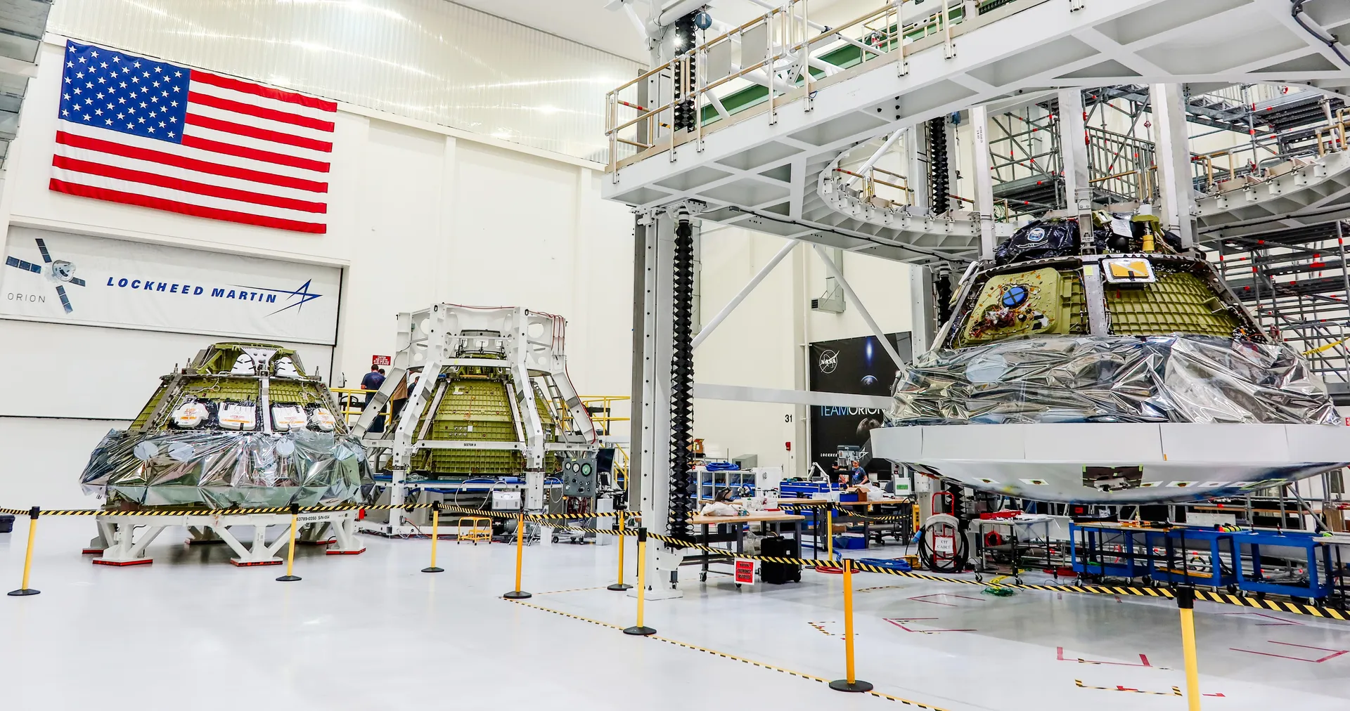 Three Orion crew modules at different stages of assembly stand side by side in a bright high bay under a large U.S. flag.