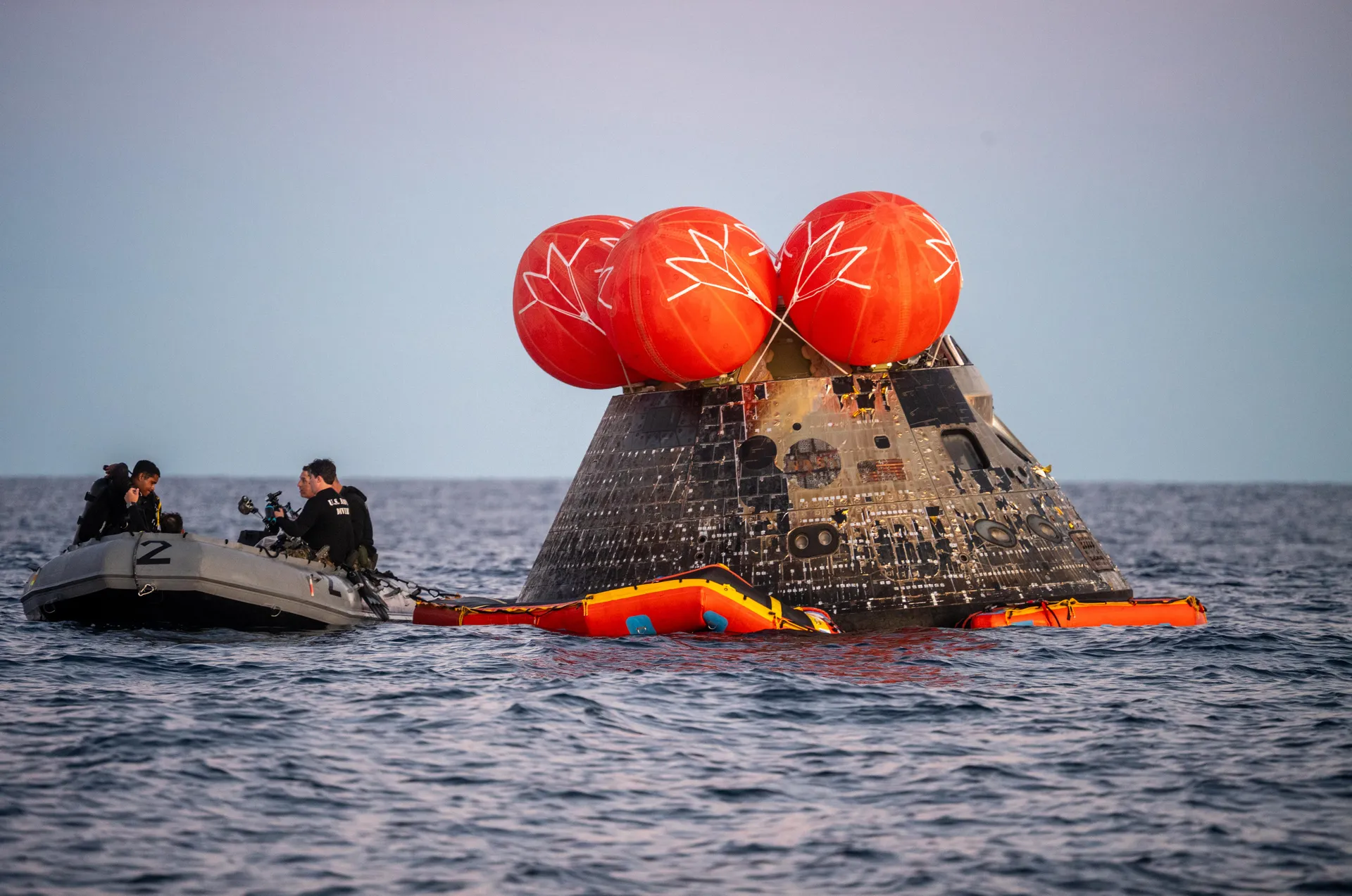 Orion floats upright in the Pacific with five orange uprighting bags on top, as Navy divers in an inflatable boat approach.
