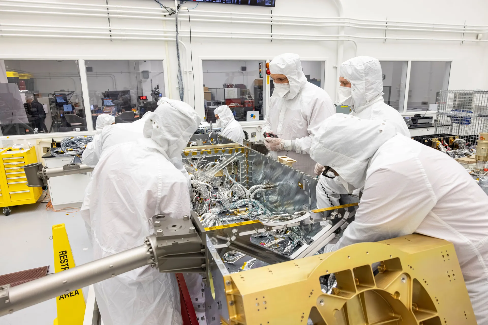 Engineers in white clean-room suits lean over an open metal frame threaded with silver cables.